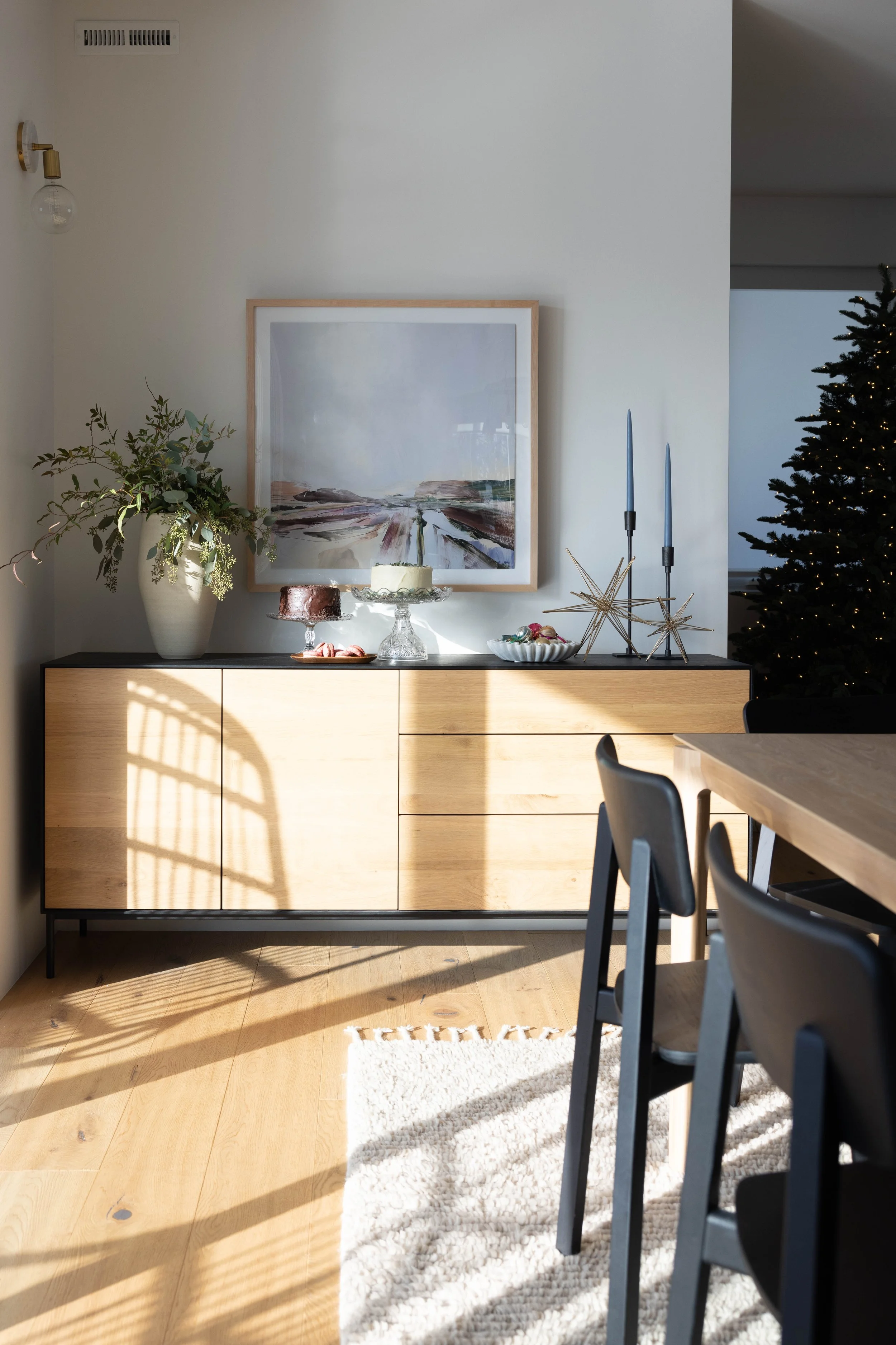 Sunlit dining area with a wood sideboard, framed art, and a black table.