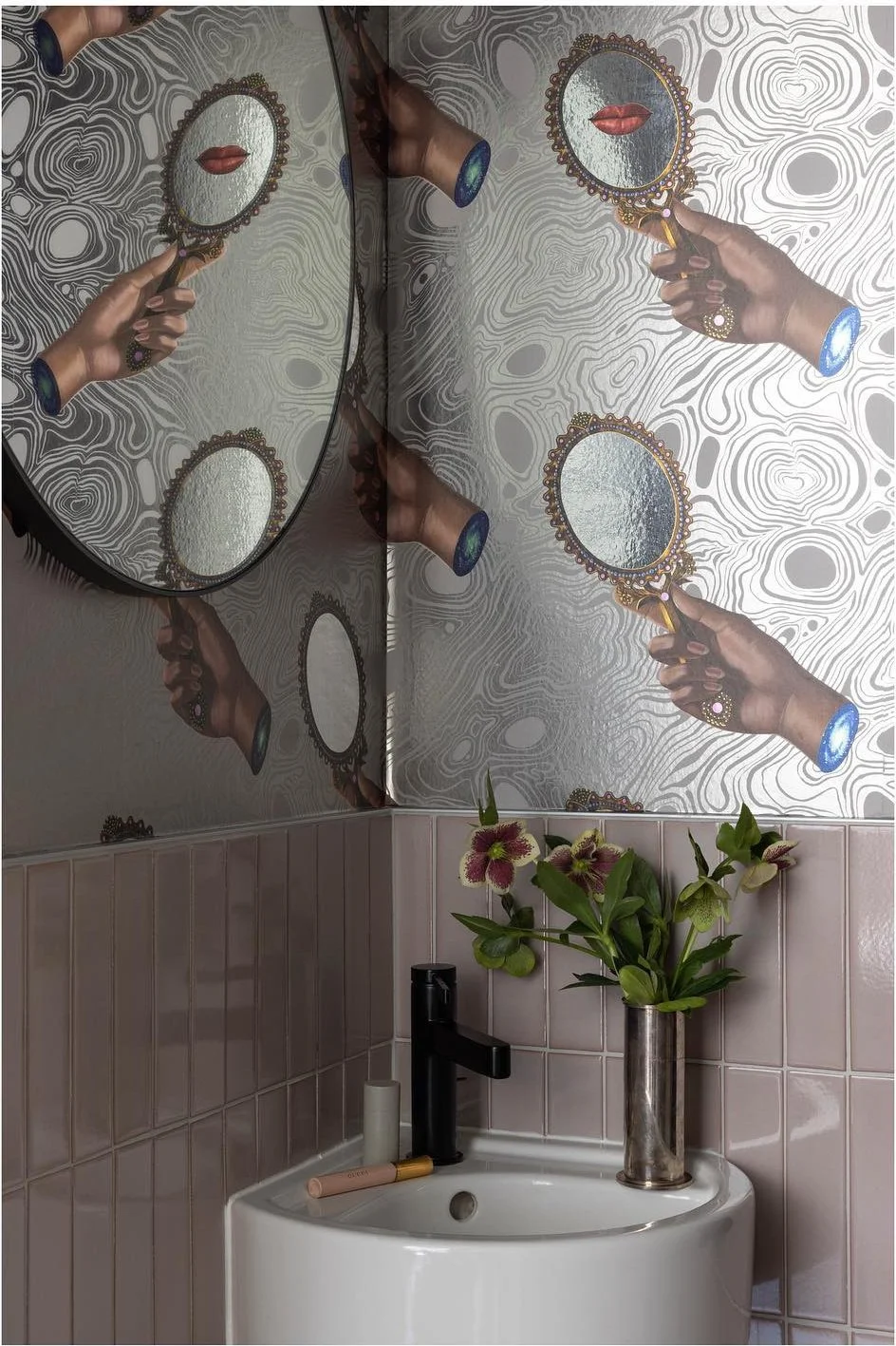Powder room with pink tile and wallpaper depicting hands holding mirrors above a small white sink.