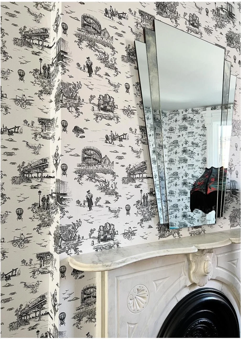 Shaped mirror above an ornate fireplace on illustrated wallpaper