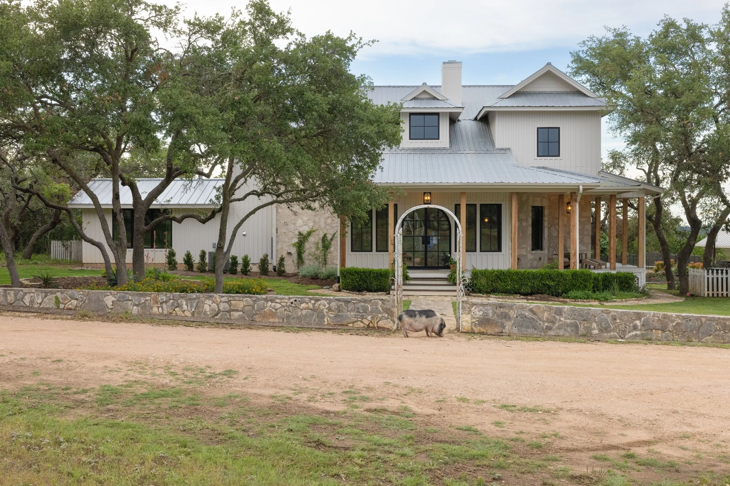 White house with a covered porch set behind trees and a stone wall.