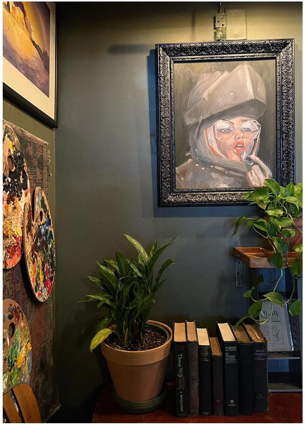 Framed portrait above plants and stacked books against a dark wall