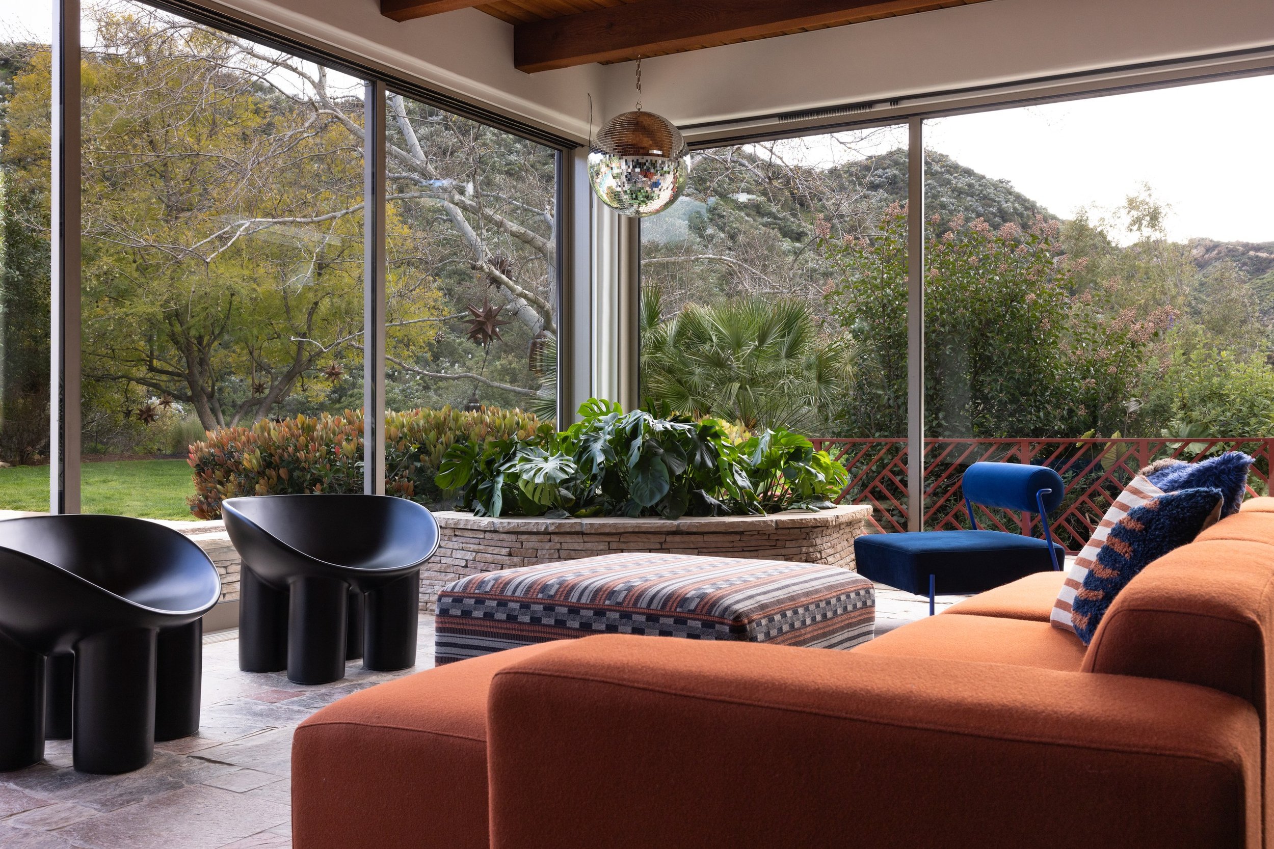 Sunroom with an orange sofa, patterned upholstered ottoman, black sculptural chairs, exposed timber, and large windows overlooking the hills.