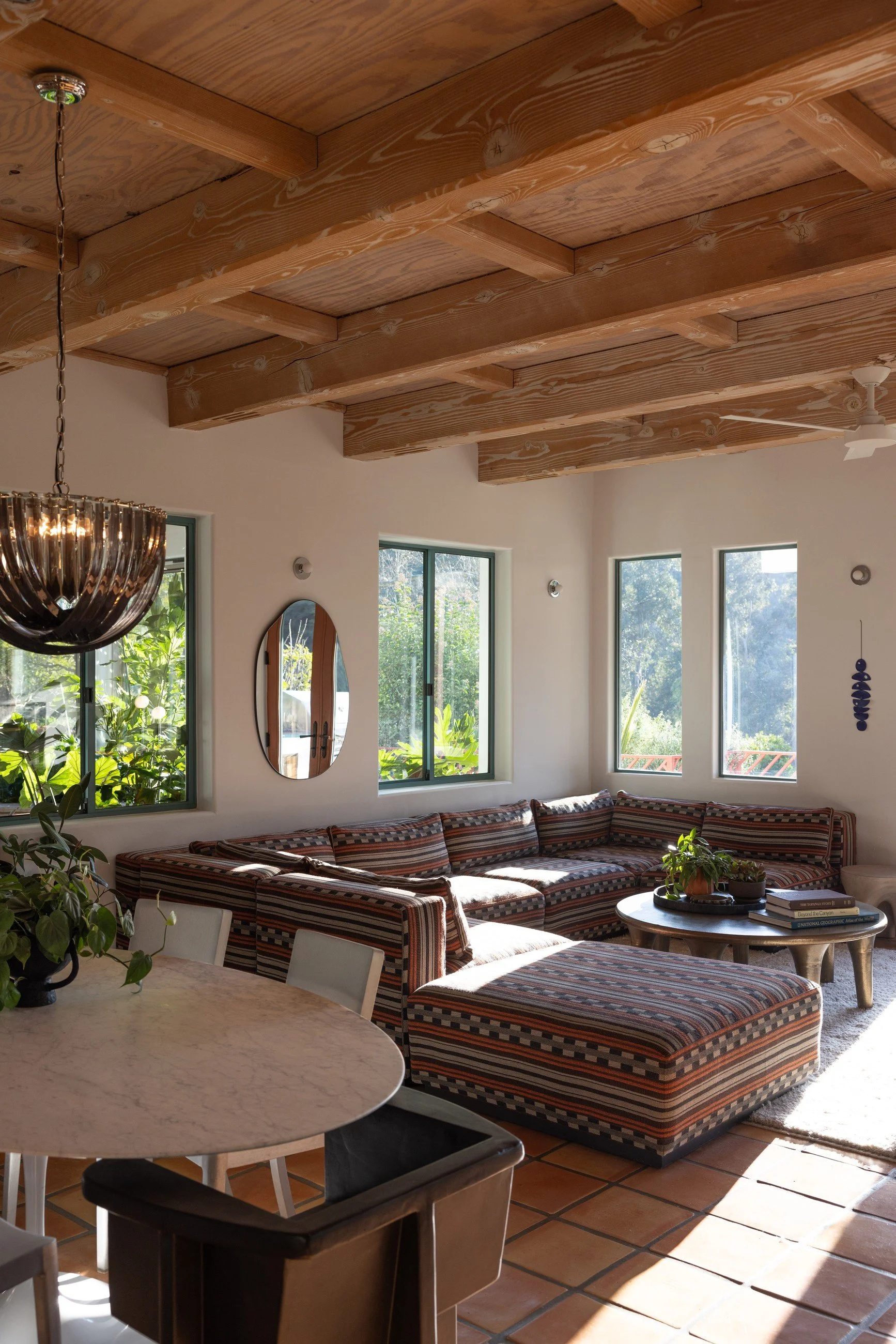 Kitchen and living room with a round white table, dark wood stools, patterned upholstered seating, Saltillo tile, and exposed beams.