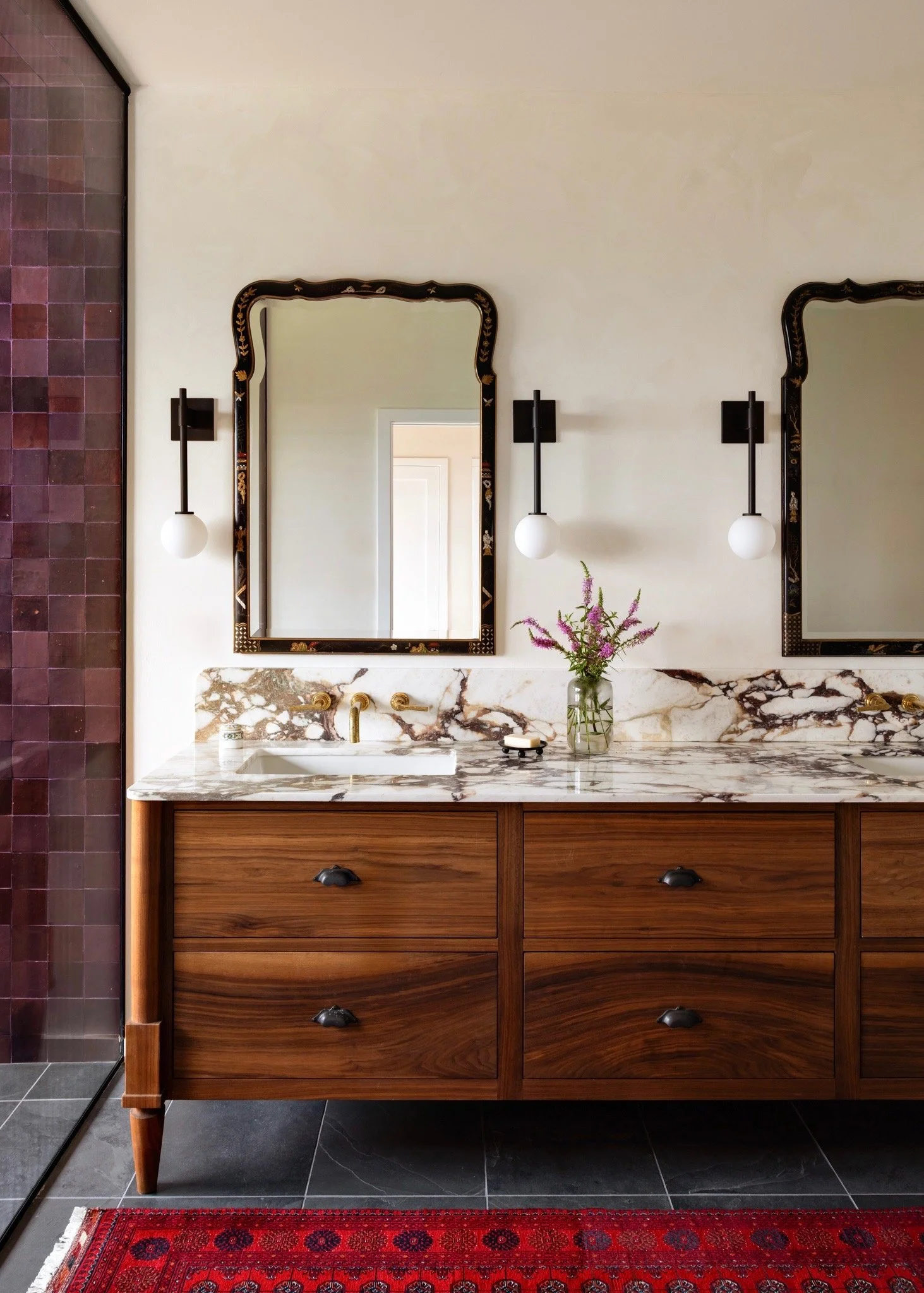 Walnut bathroom vanity with a marble counter, ornate mirrors, and globe sconces.