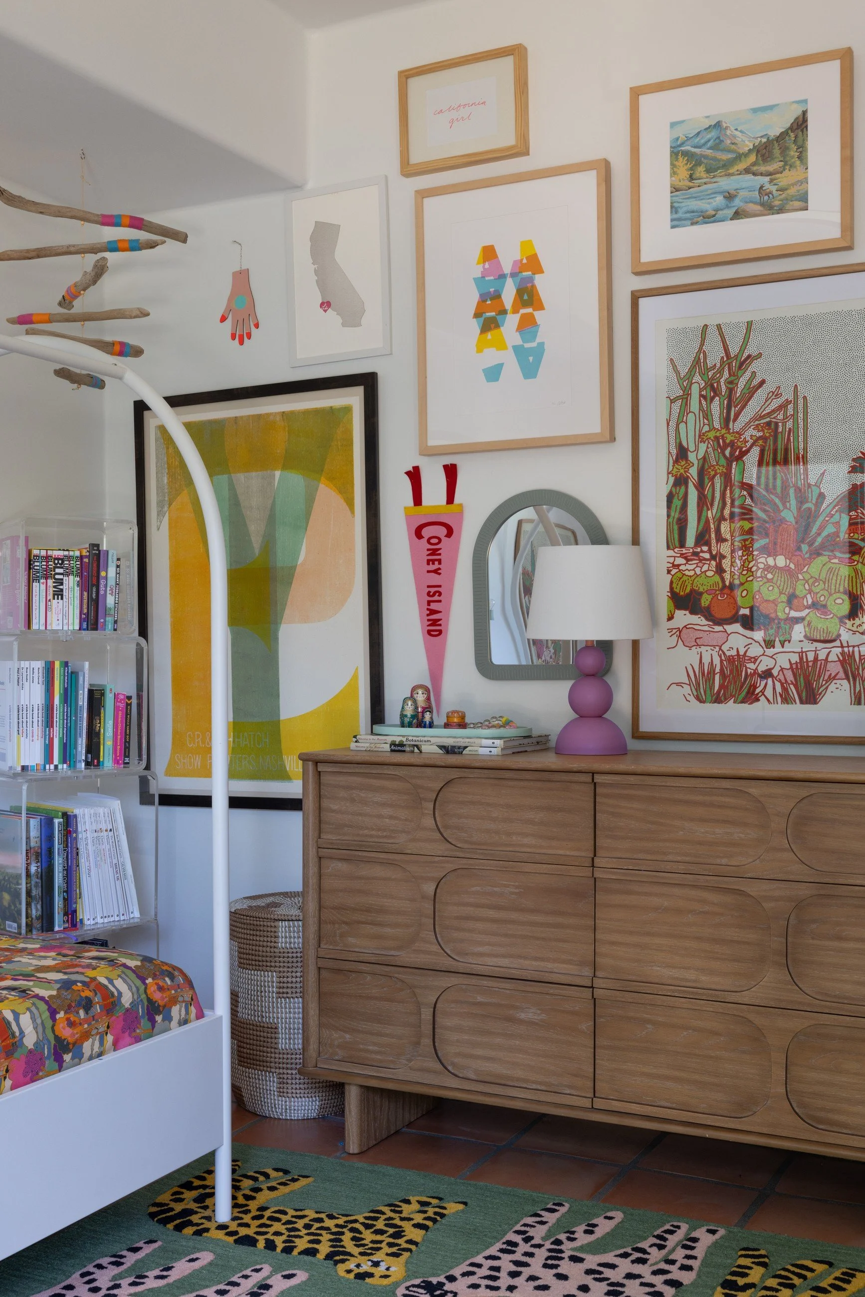 Bedroom vignette with a wood dresser, colorful framed artwork, floral bed textiles, and a patterned green rug.