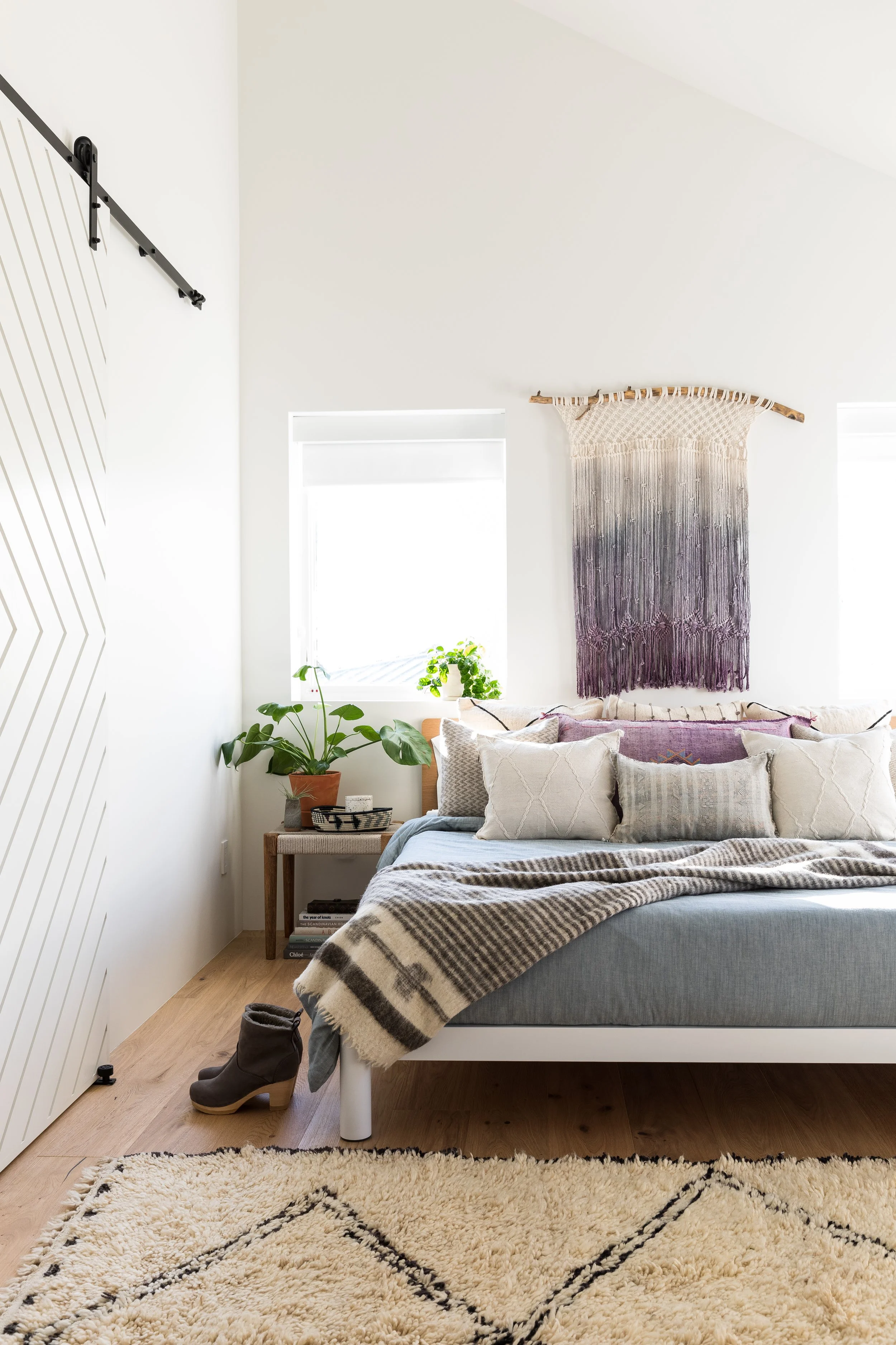 Bright bedroom with layered bedding, woven wall hanging, and patterned rug.