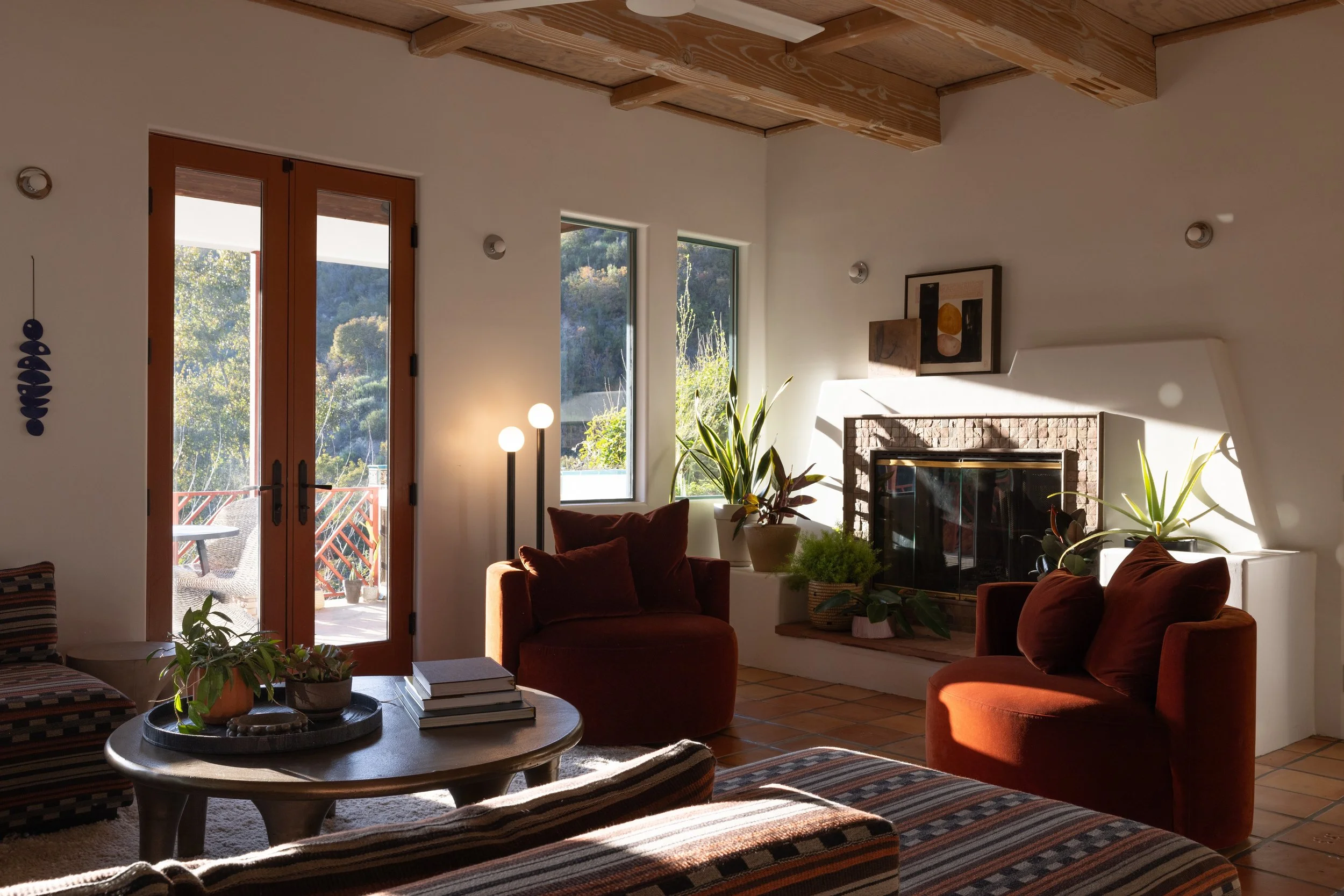 Sunlit living room with rust-colored chairs, a tiled fireplace, wood-beam ceiling and garden views.