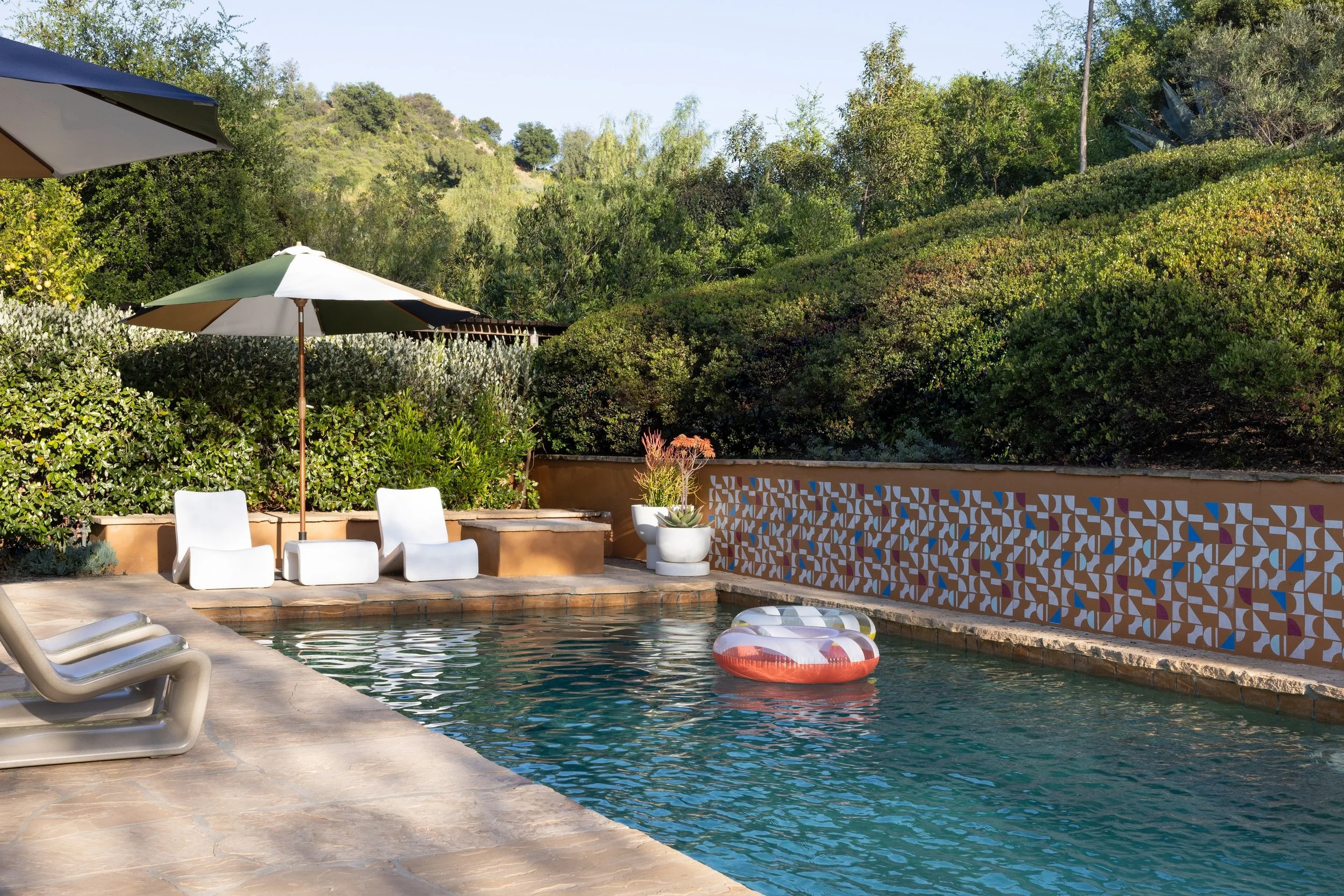 Swimming pool with a patterned tile waterline, white loungers, a striped umbrella, and dense California hillside planting.