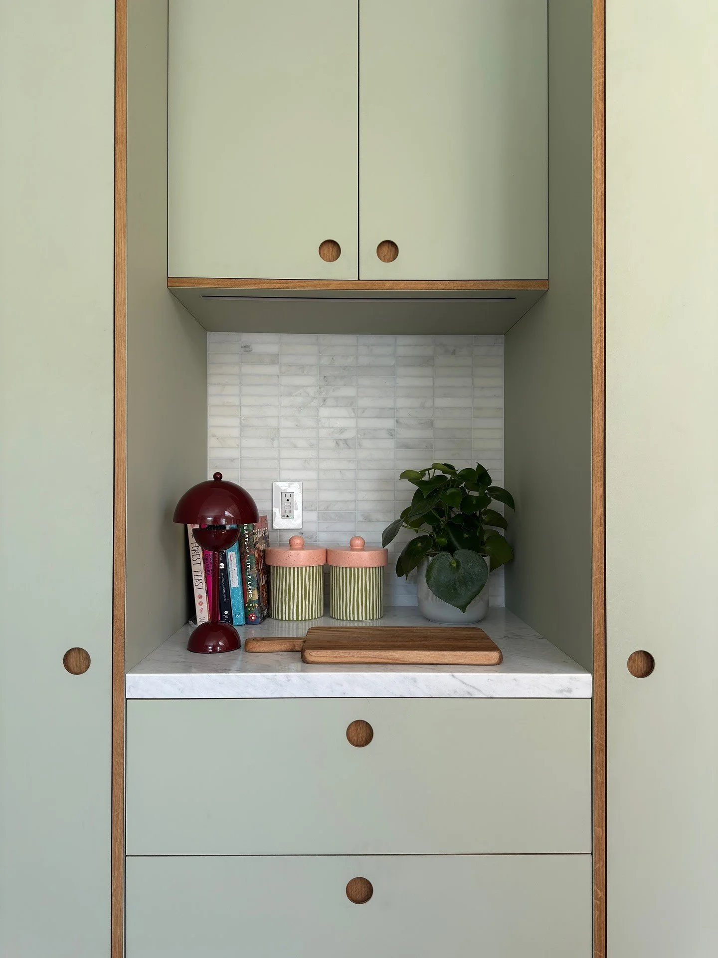 Pale green pantry cabinetry framing a styled counter with a red lamp
