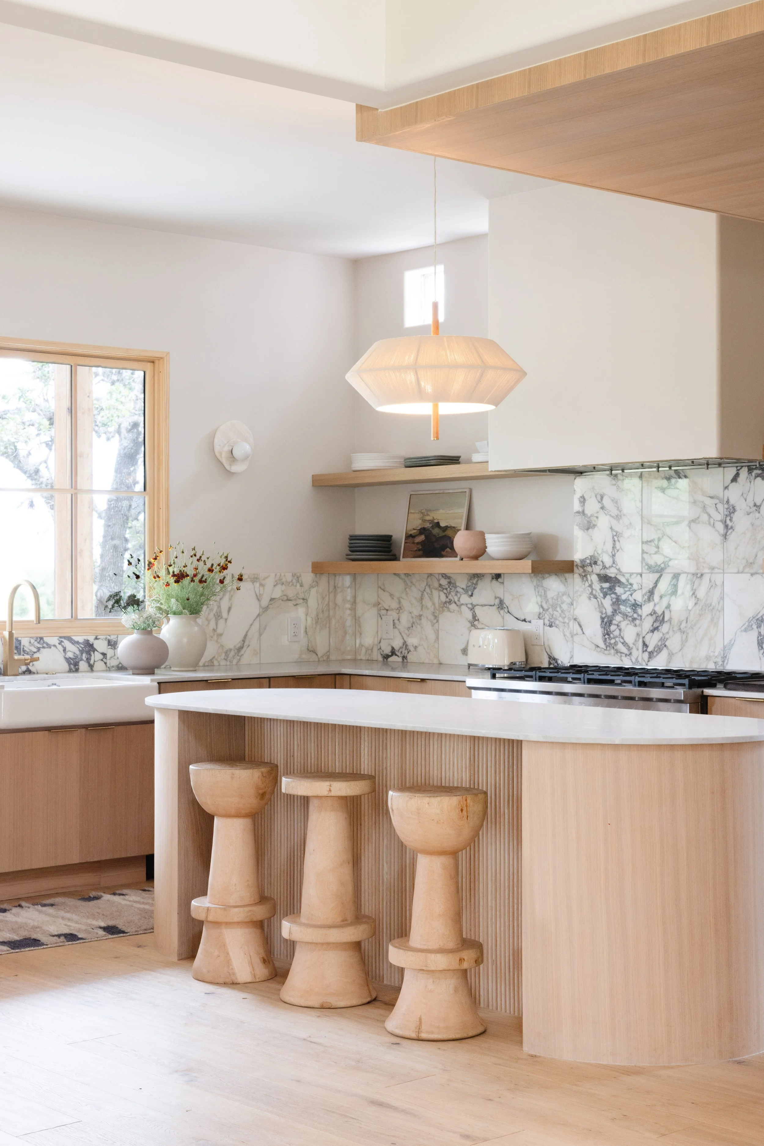 Light wood kitchen with marble backsplash, curved island, and sculptural stools.