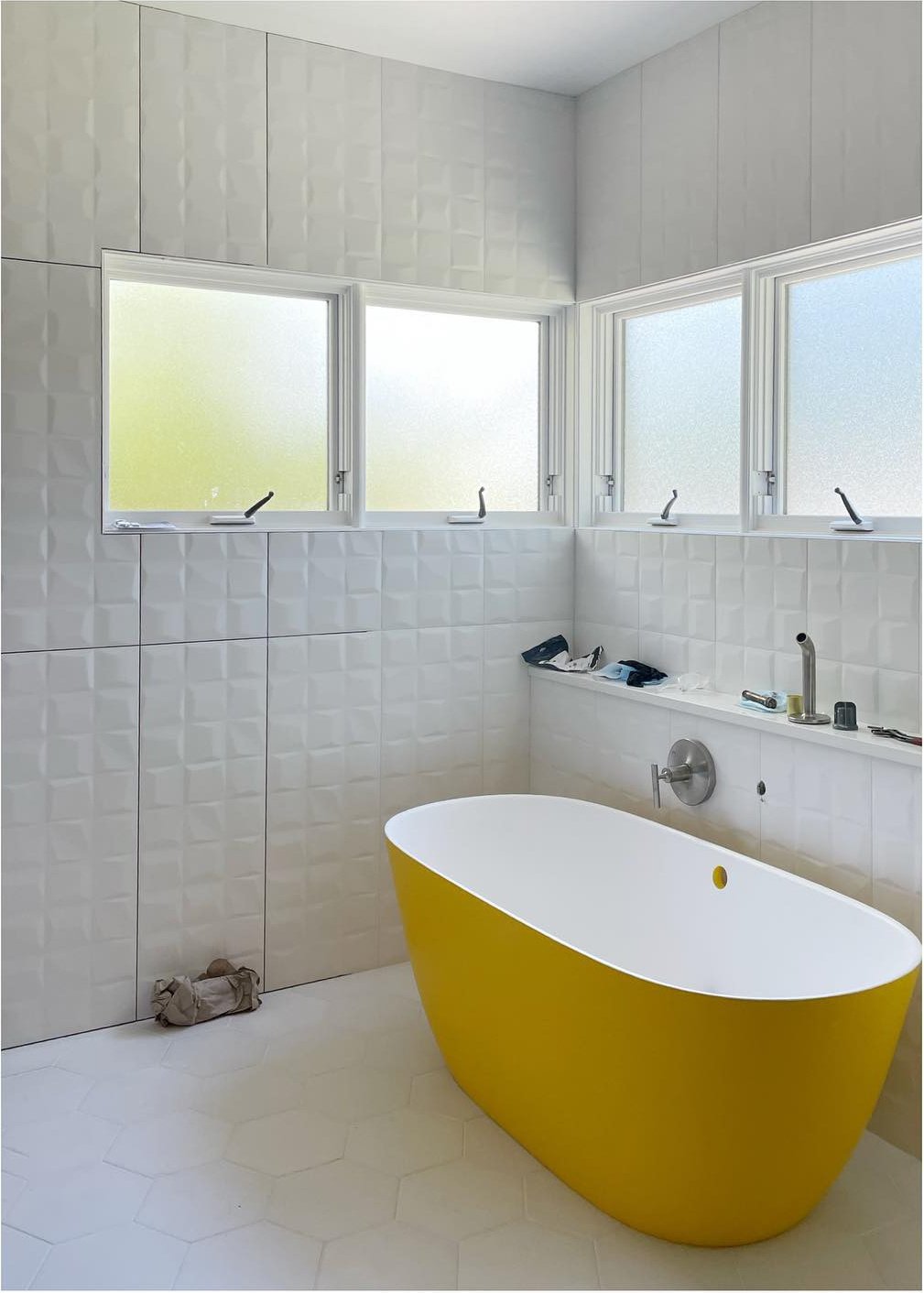 Yellow freestanding bathtub beneath bright windows and pale vertical wall tiles