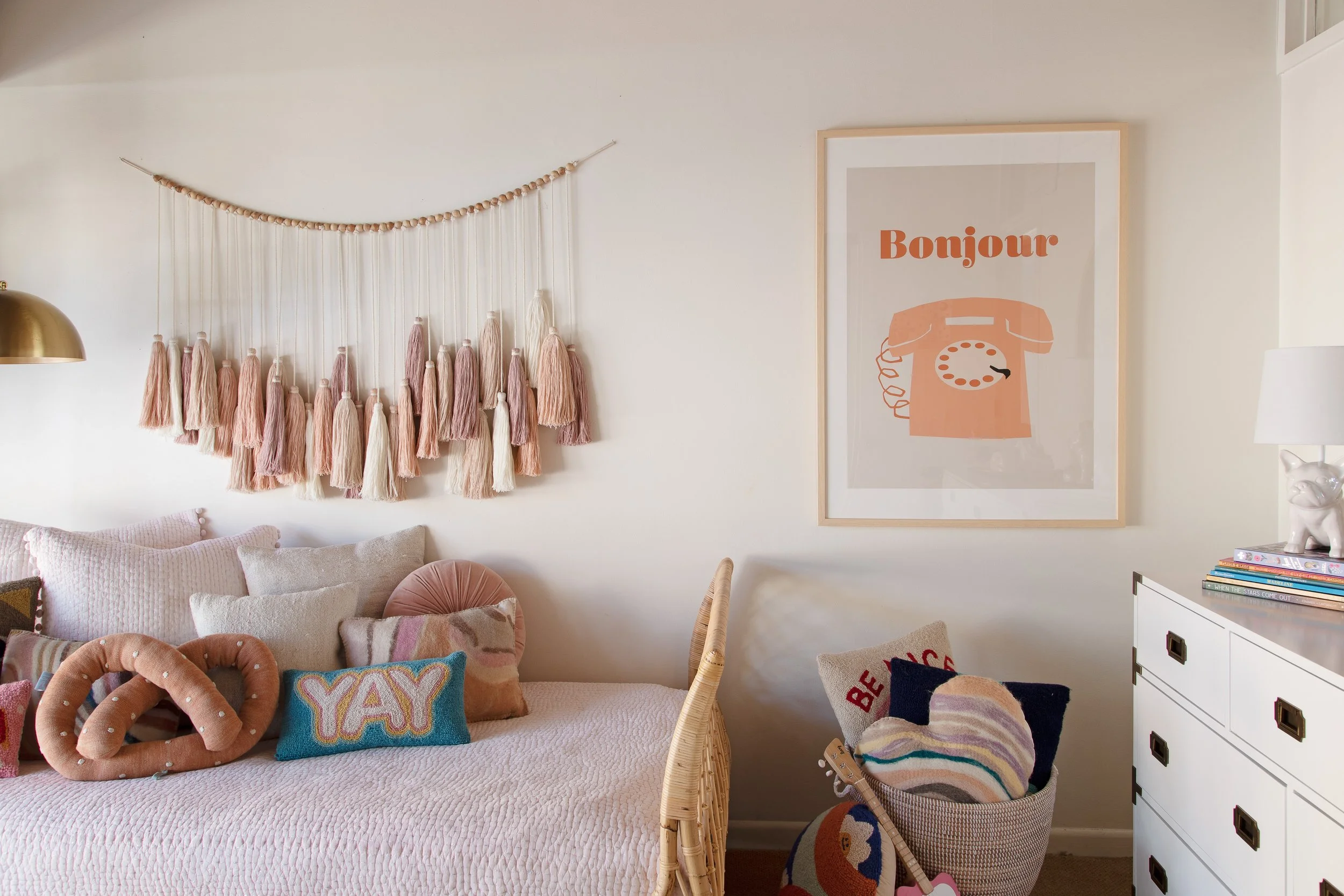 Child’s bedroom with a pink bed, tassel wall hanging, and framed art.