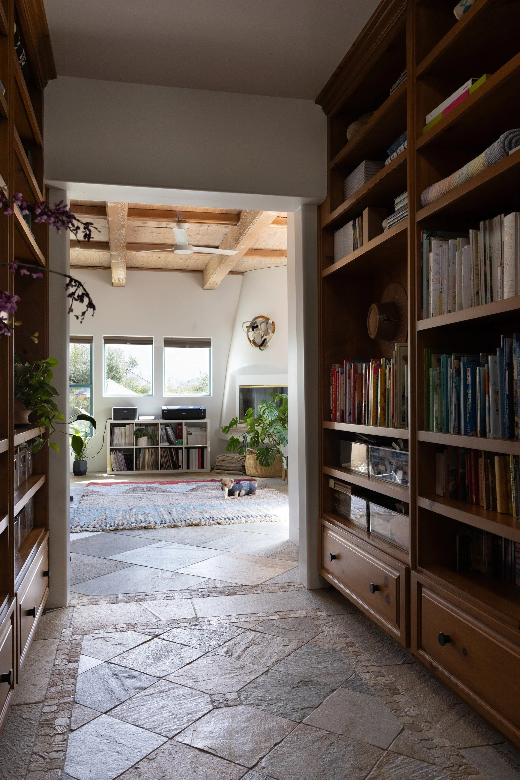 Wood-lined library passage with full-height bookshelves, stone flooring, and a view through to the bright living room.