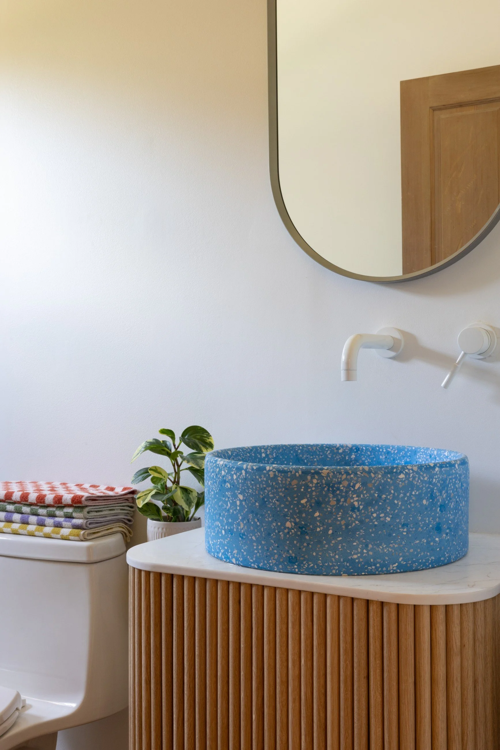 Blue stone vessel sink on a ribbed wood vanity beneath a rounded mirror, with a white wall-mounted faucet and potted plant.