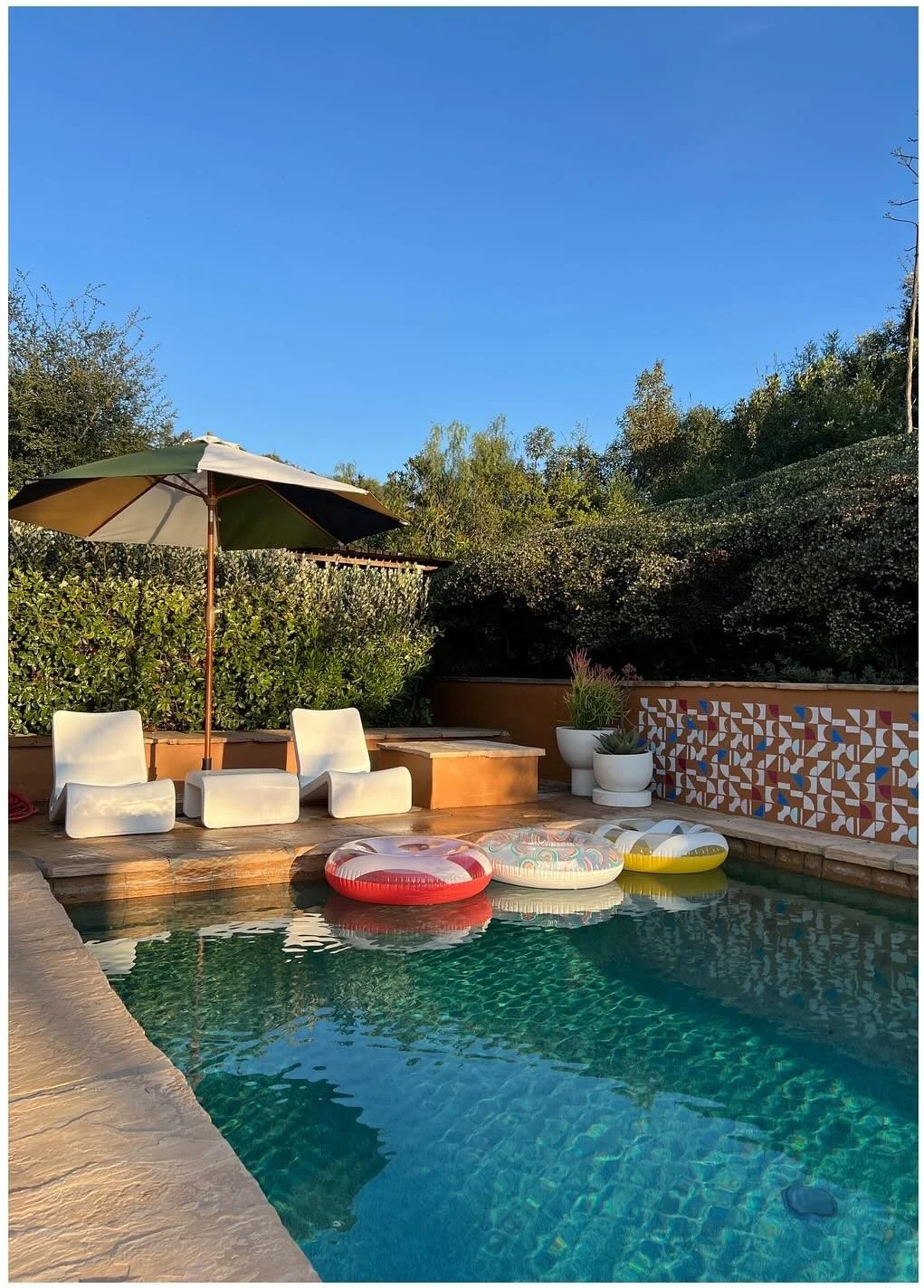 Poolside white chairs beneath a striped umbrella beside patterned tile