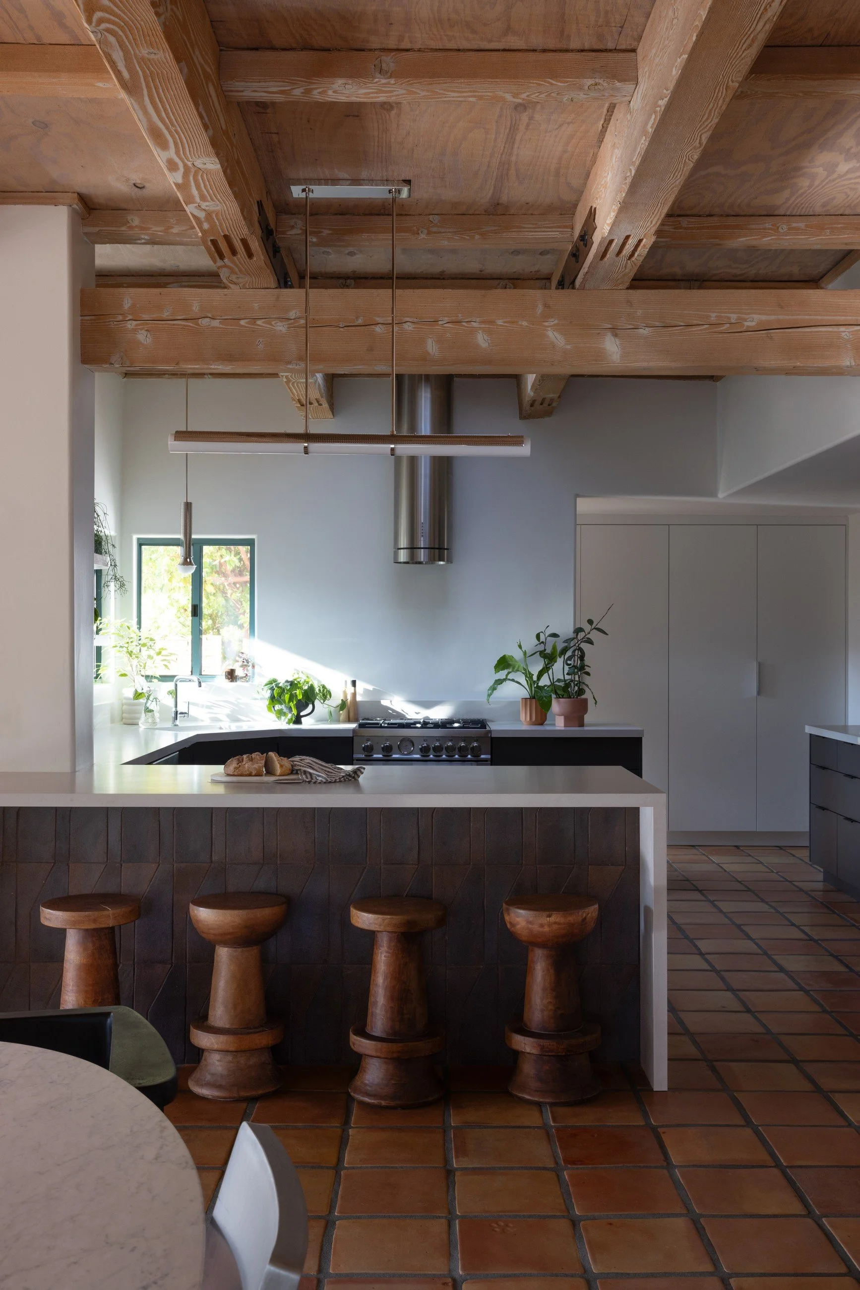 Kitchen island with dark wood cladding and turned wood stools beneath a metal range hood and exposed timber ceiling.