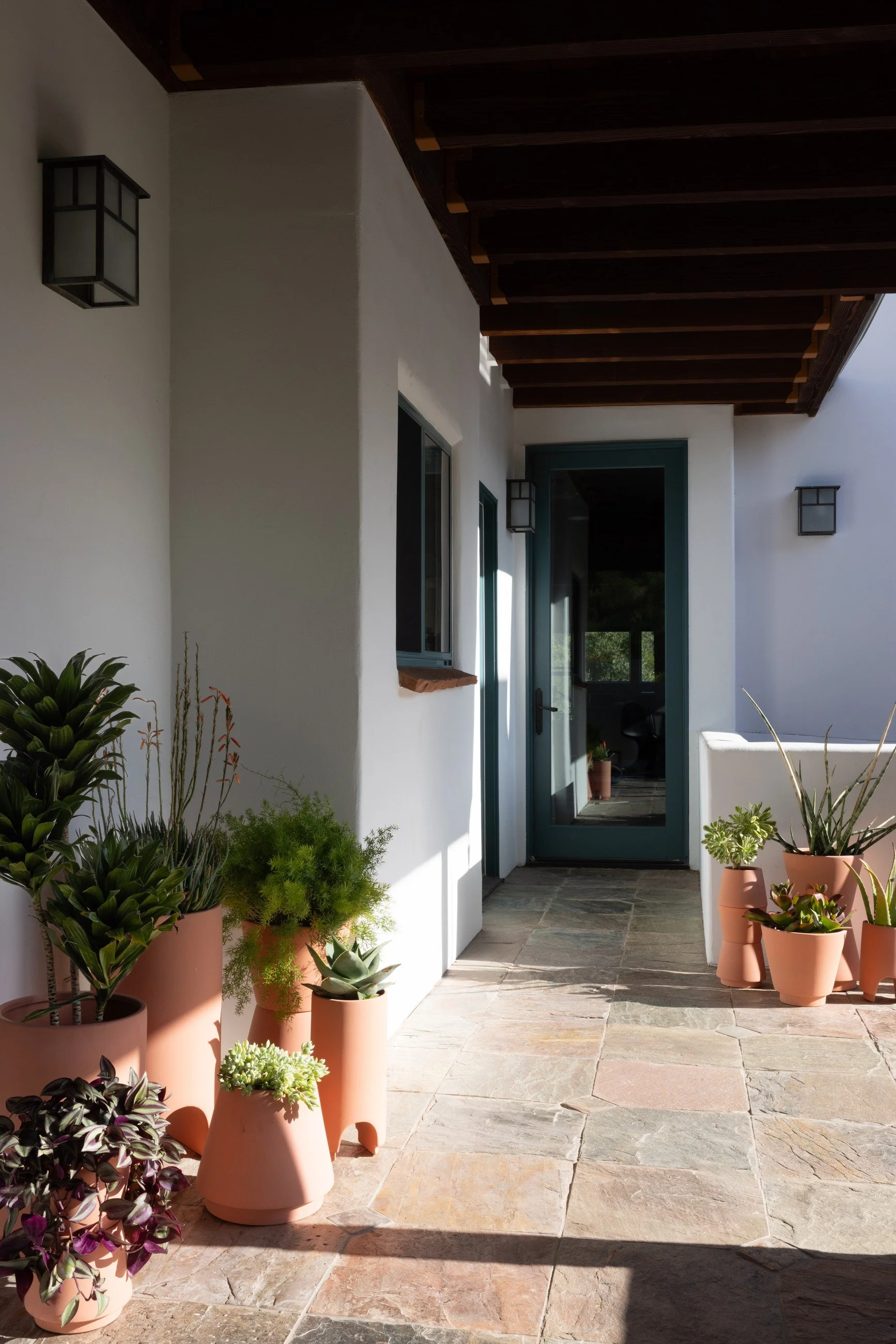 Shaded porch passage with dark overhead beams, a row of terracotta planters, and an open doorway into the house.