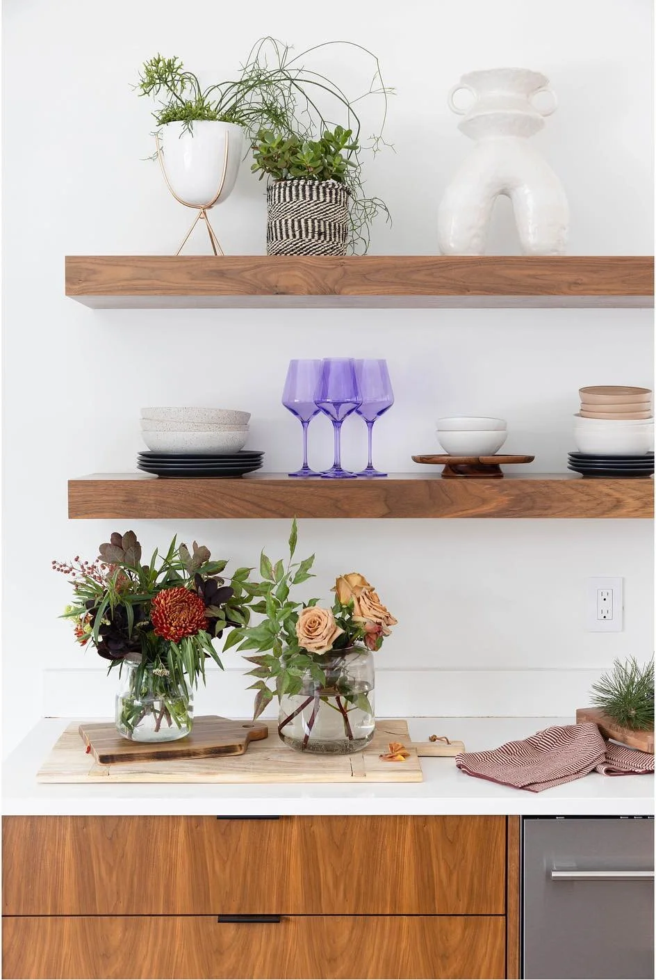 Open wood shelves displaying plants glasses and ceramics above a kitchen counter