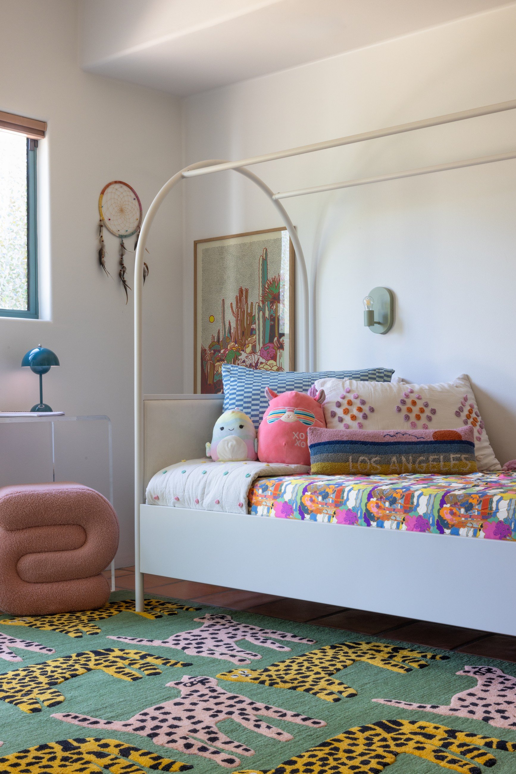 Colorful bedroom with a white canopy bed, floral bedding, patterned green rug, framed art, and a pink upholstered chair.
