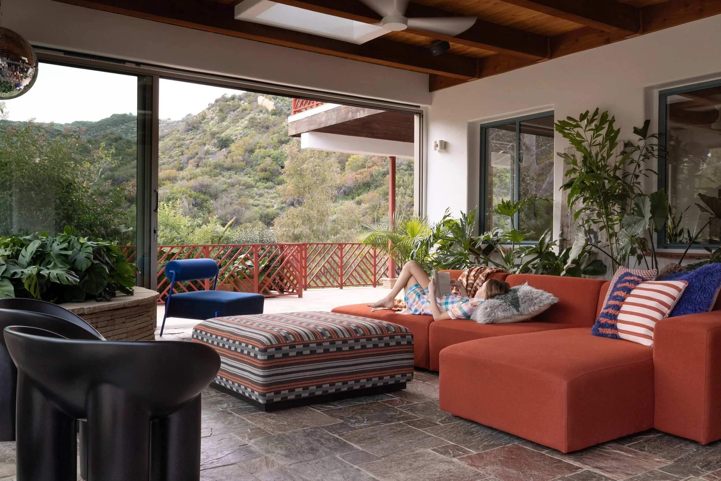 Orange sectional sofa and patterned ottoman beneath a wood-beam ceiling in a sunroom surrounded by garden views.