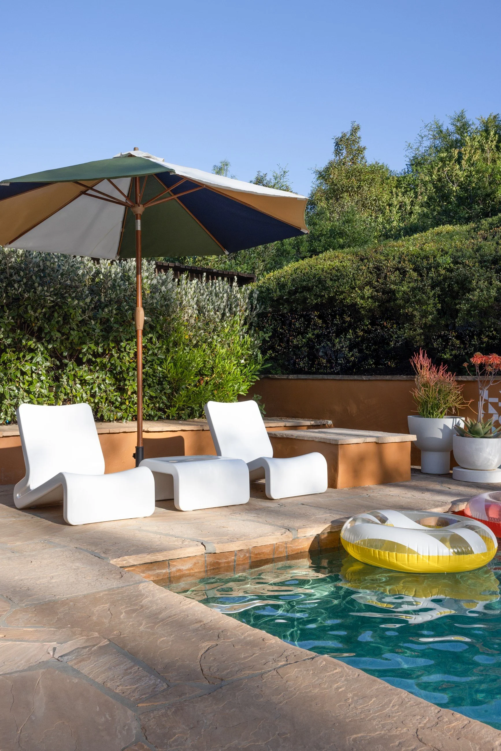 Poolside patio with a striped umbrella, white lounge chairs, warm stone paving, and a planted hillside.