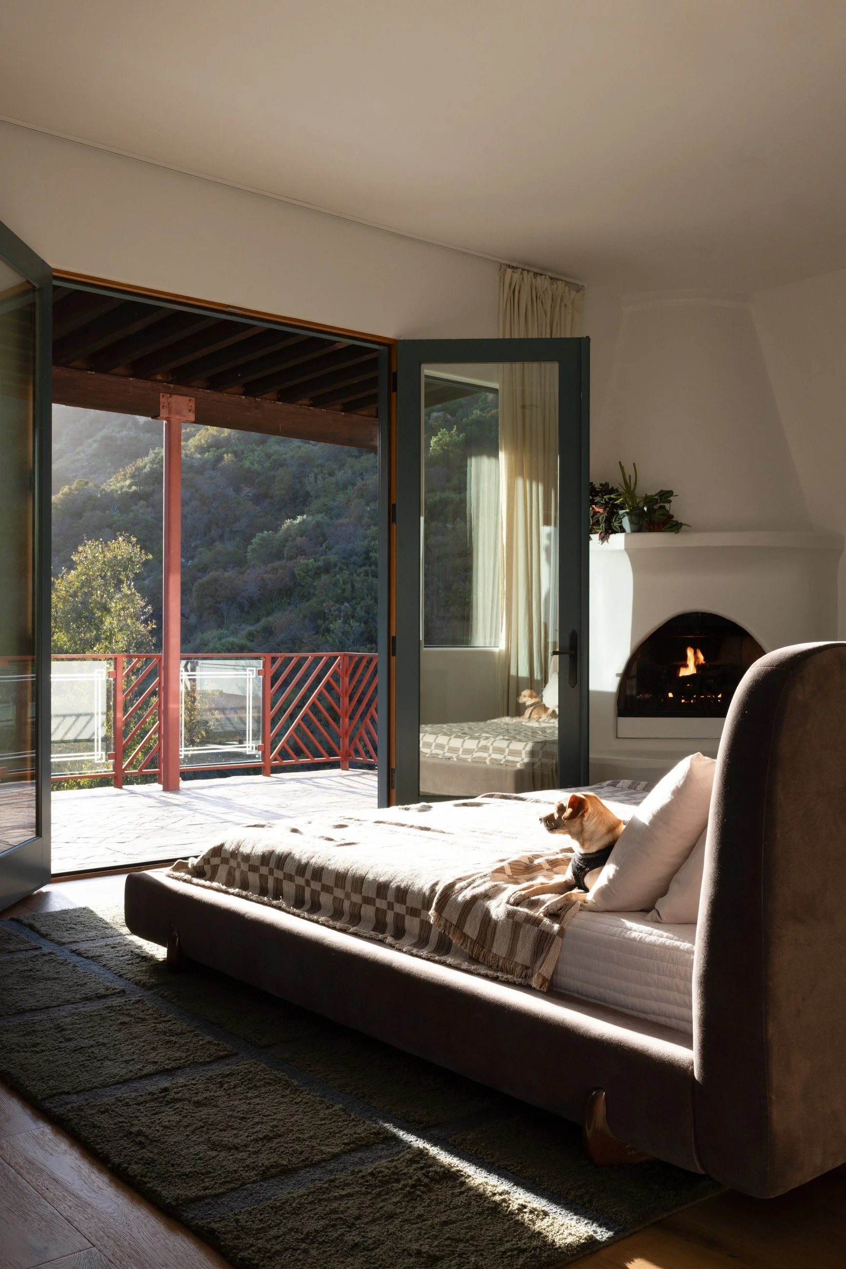 Bedroom with a low upholstered bed, neutral bedding, a dark rug, and open glass doors facing the California hillside.