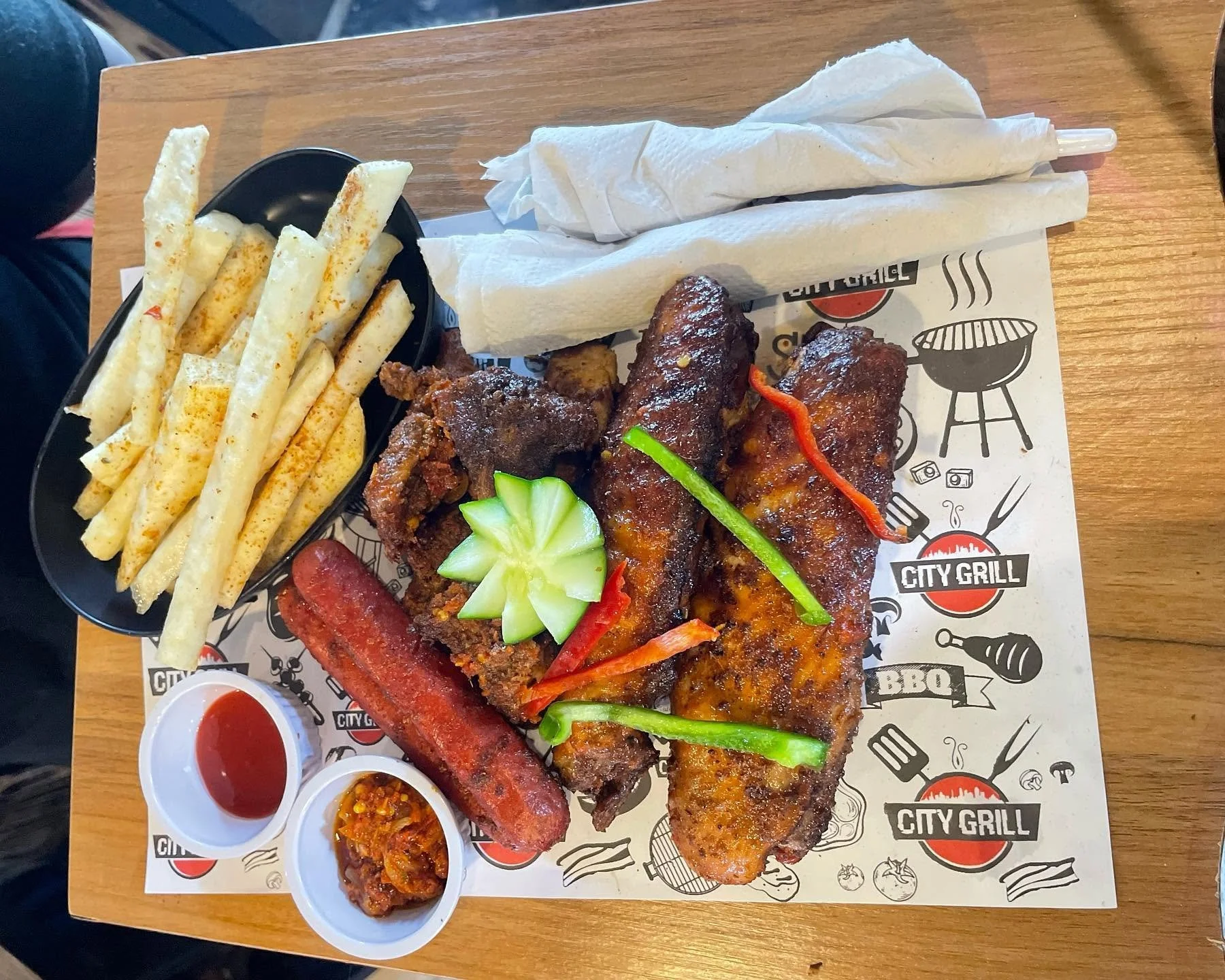 Overhead view of a barbecue platter with grilled meats, seasoned fries, sliced peppers, cucumber garnish, dipping sauces, and wrapped utensils.