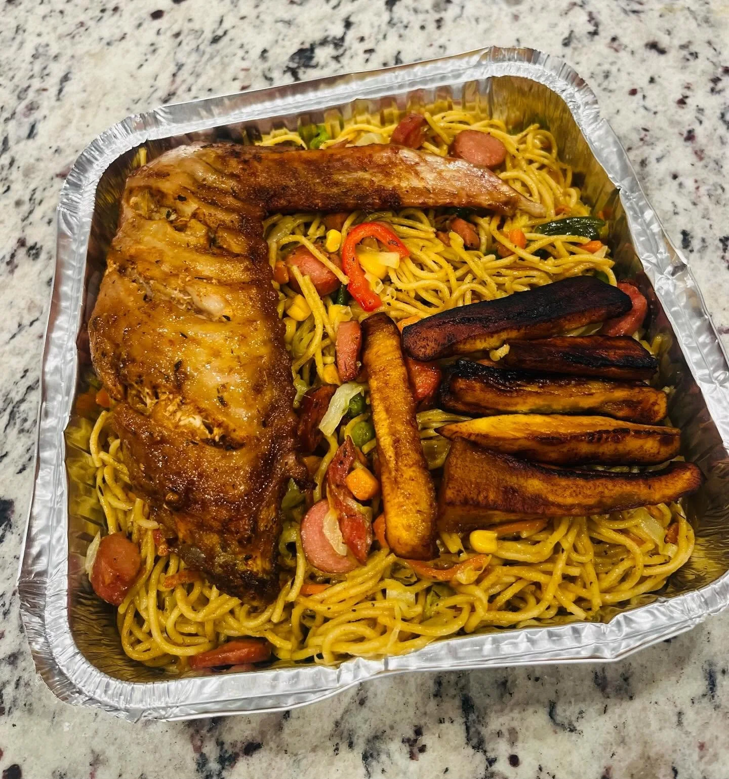 Aluminum takeout tray filled with stir-fried noodles, roasted chicken, sausage, fried plantains, and mixed vegetables