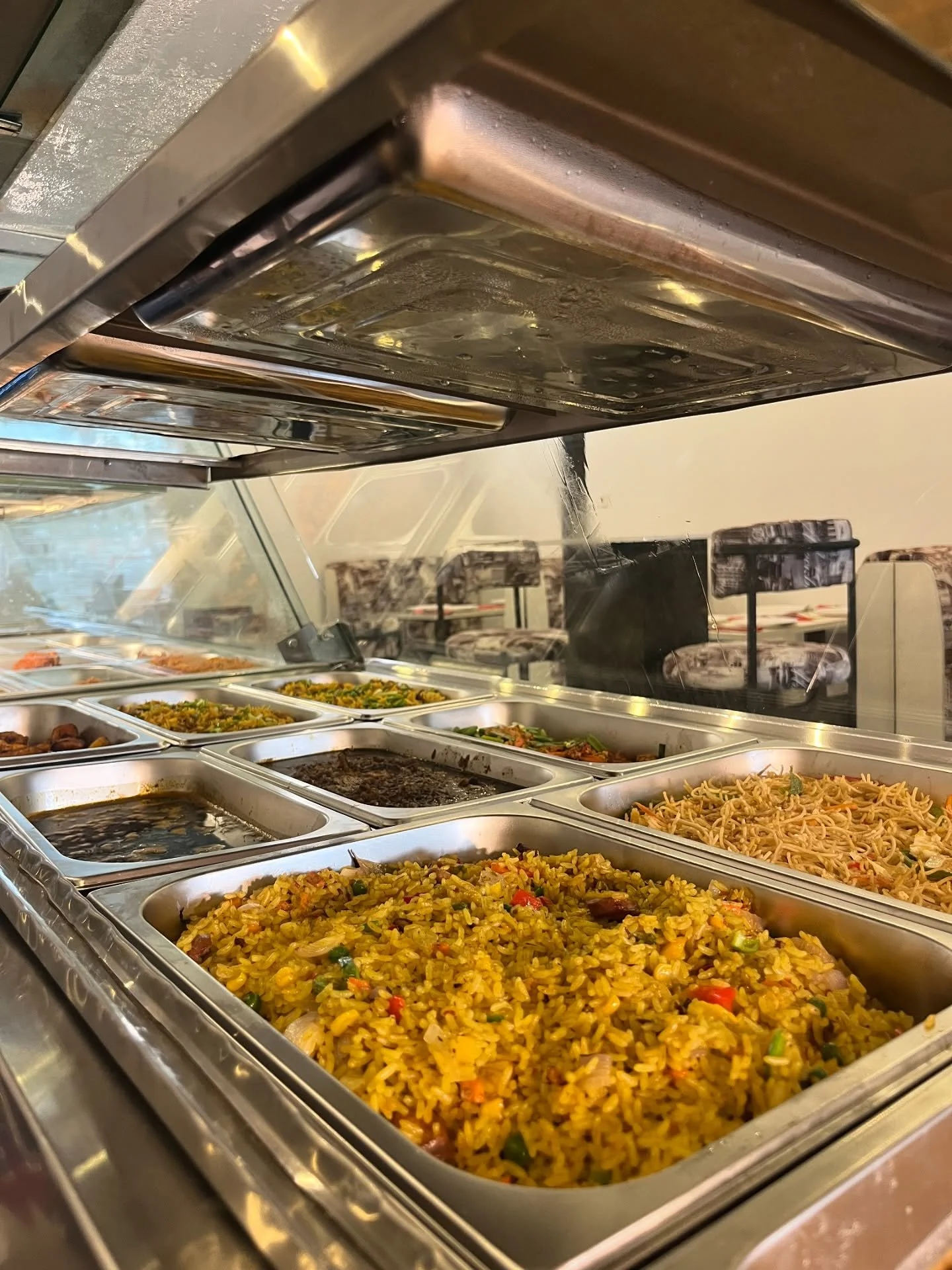 Stainless steel buffet pans filled with seasoned rice, noodles, vegetables, beans, and other prepared dishes under a heated food display.