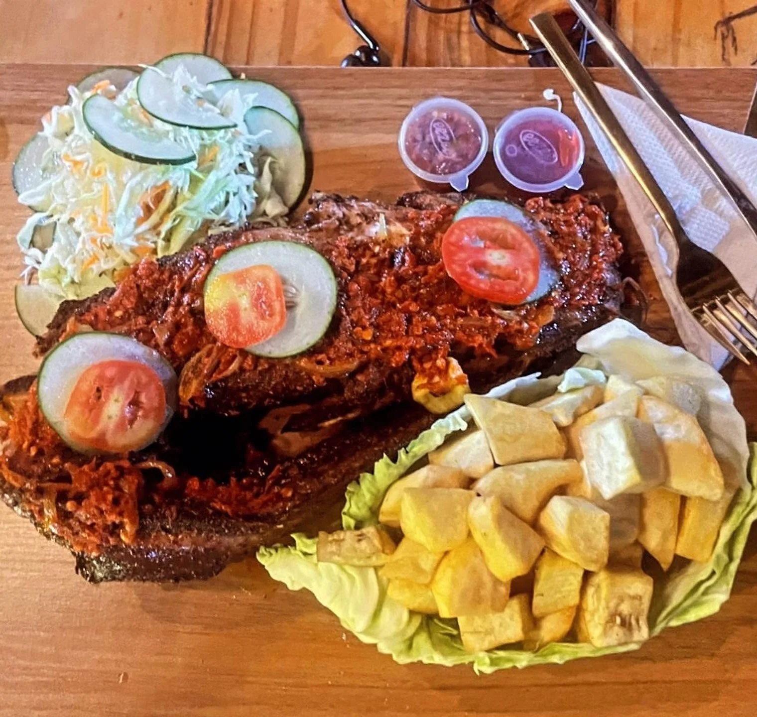 Platter of saucy grilled meat topped with tomatoes and cucumbers, cabbage salad, fried cassava, and dipping sauces