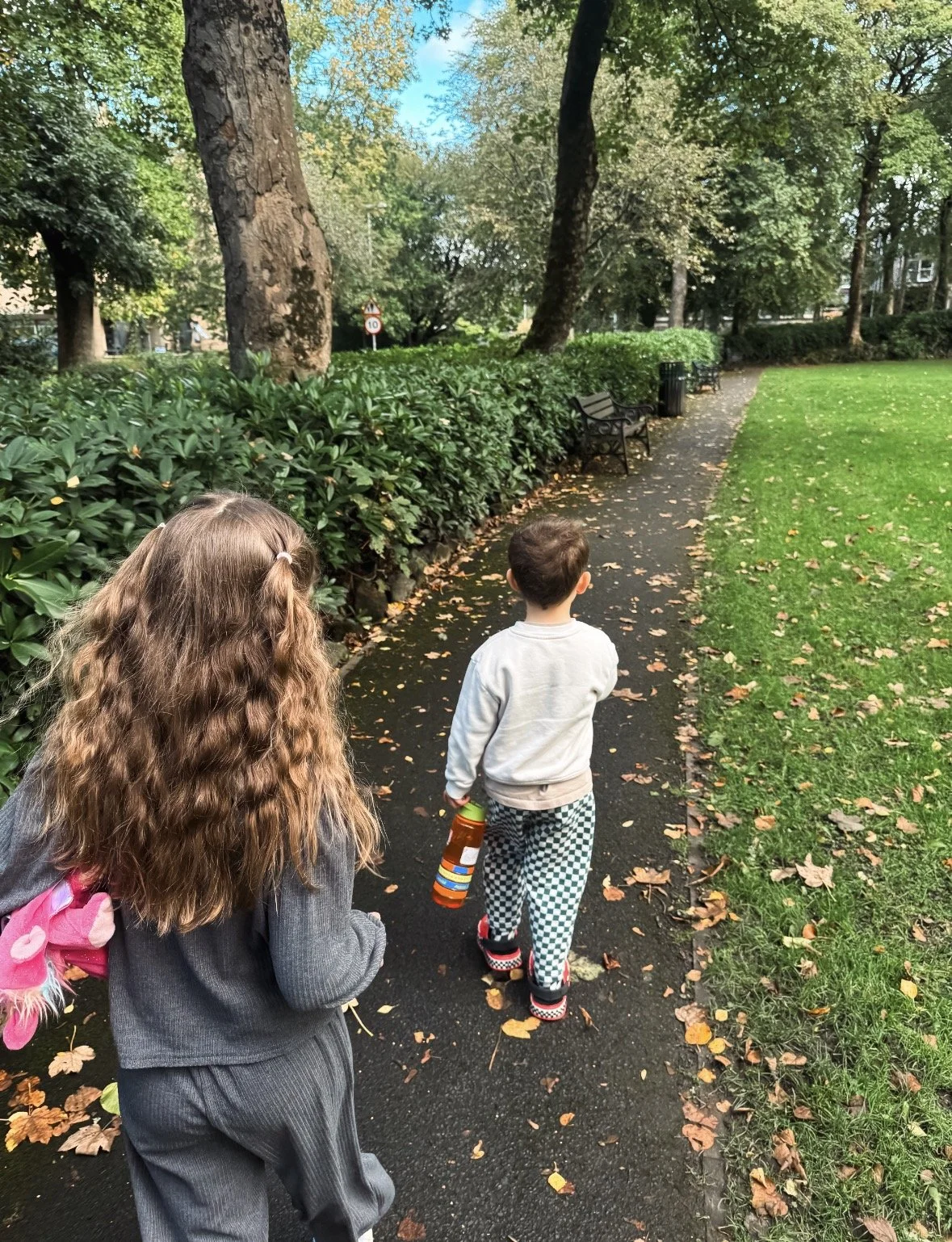 Two children walking along a tree-lined park path with benches and green lawns.