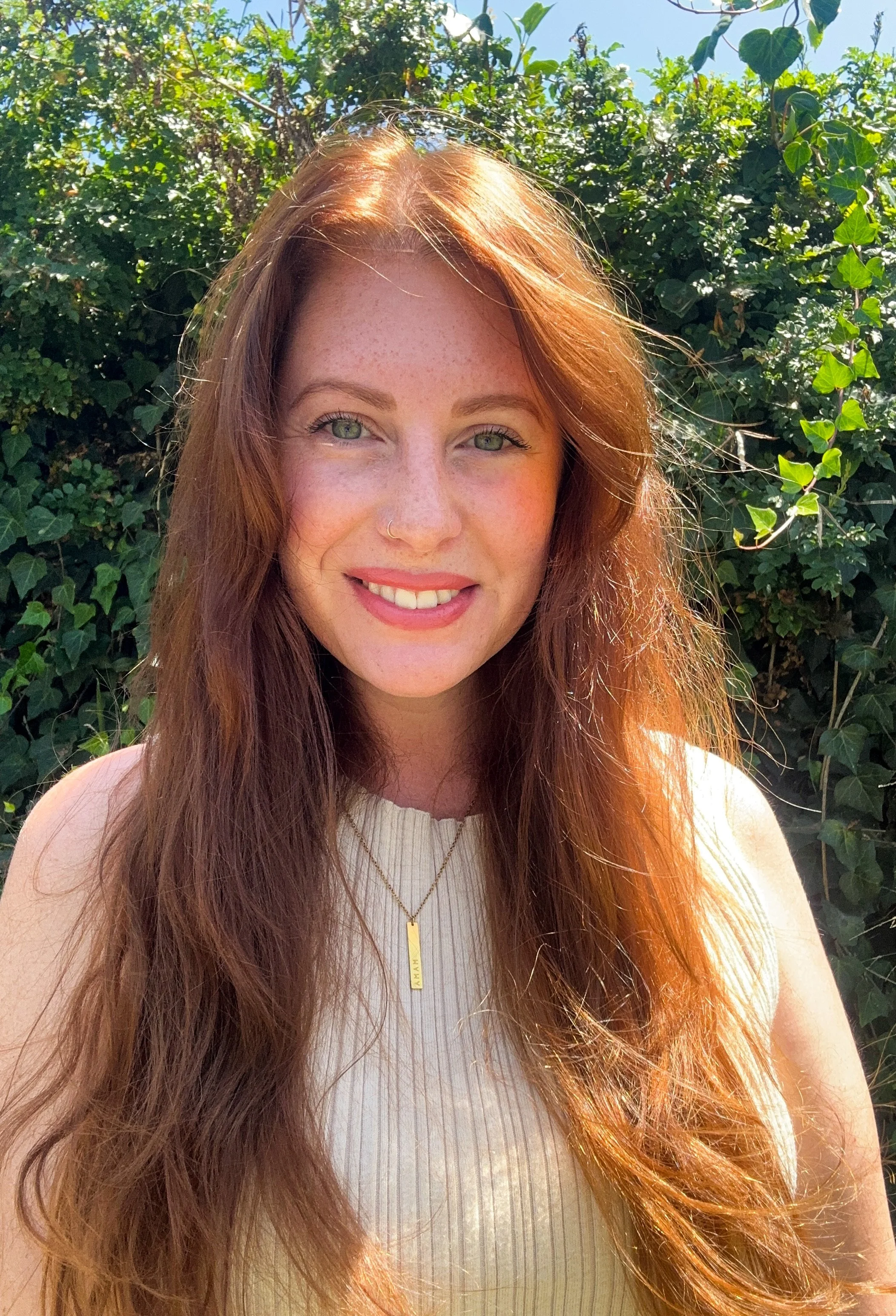 Smiling red-haired woman taking a close-up outdoor selfie in front of lush greenery