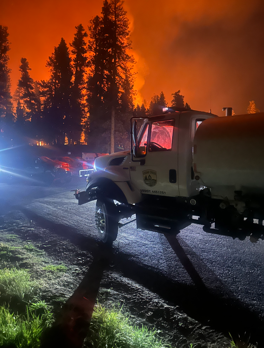 Water tanker truck parked near a forest wildfire at night, with towering pine trees silhouetted against an orange smoke-filled sky