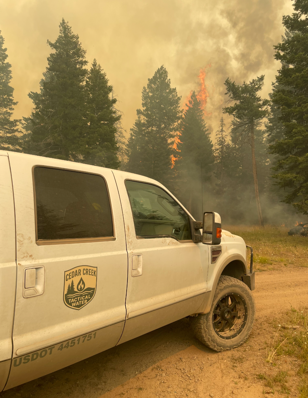 White Cedar Creek Tactical Water pickup truck parked on a dirt road as flames and heavy smoke rise through a forest during a wildfire