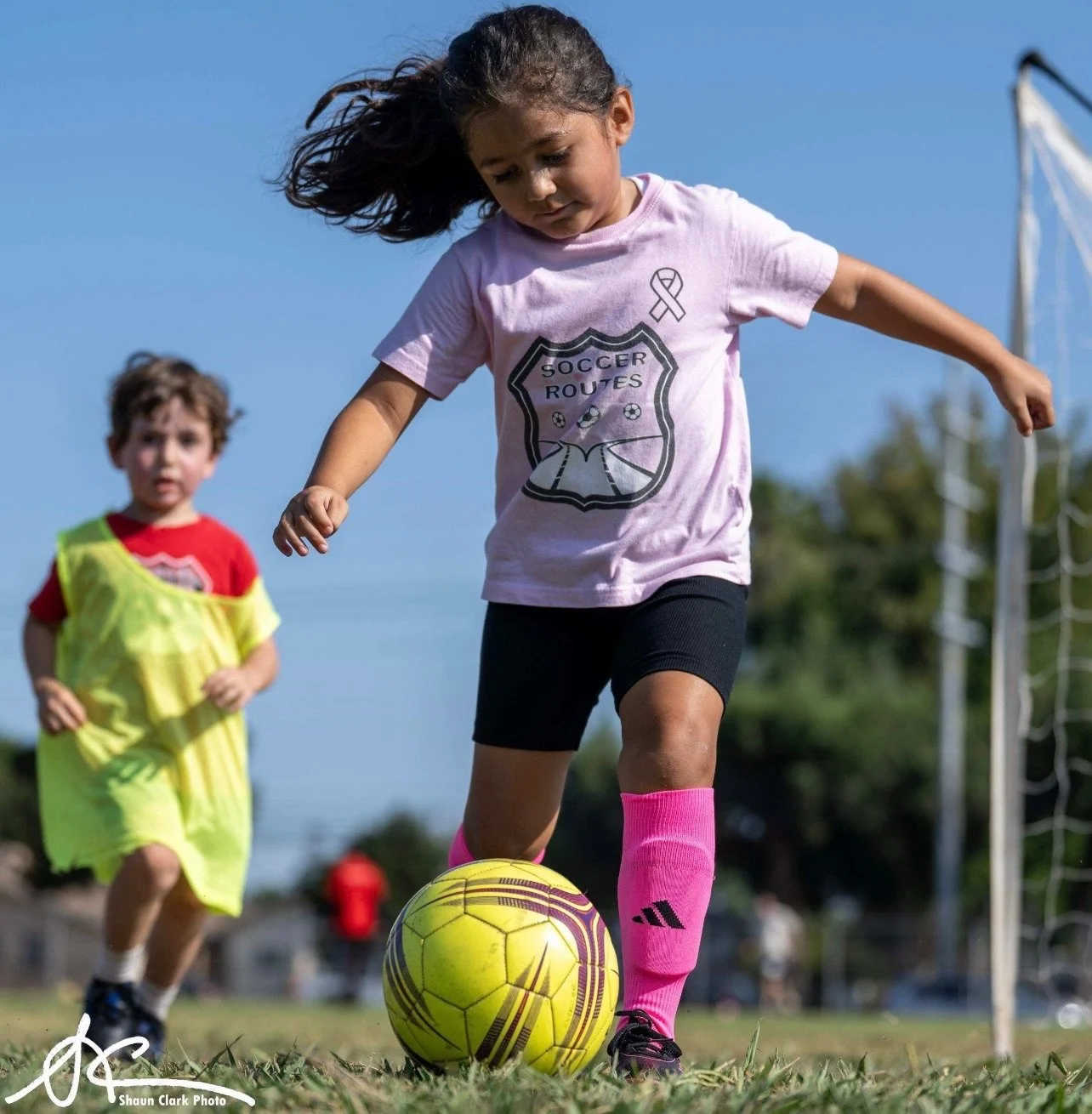 Young girl dribbling a soccer ball on a field while another child in a training bib follows behind