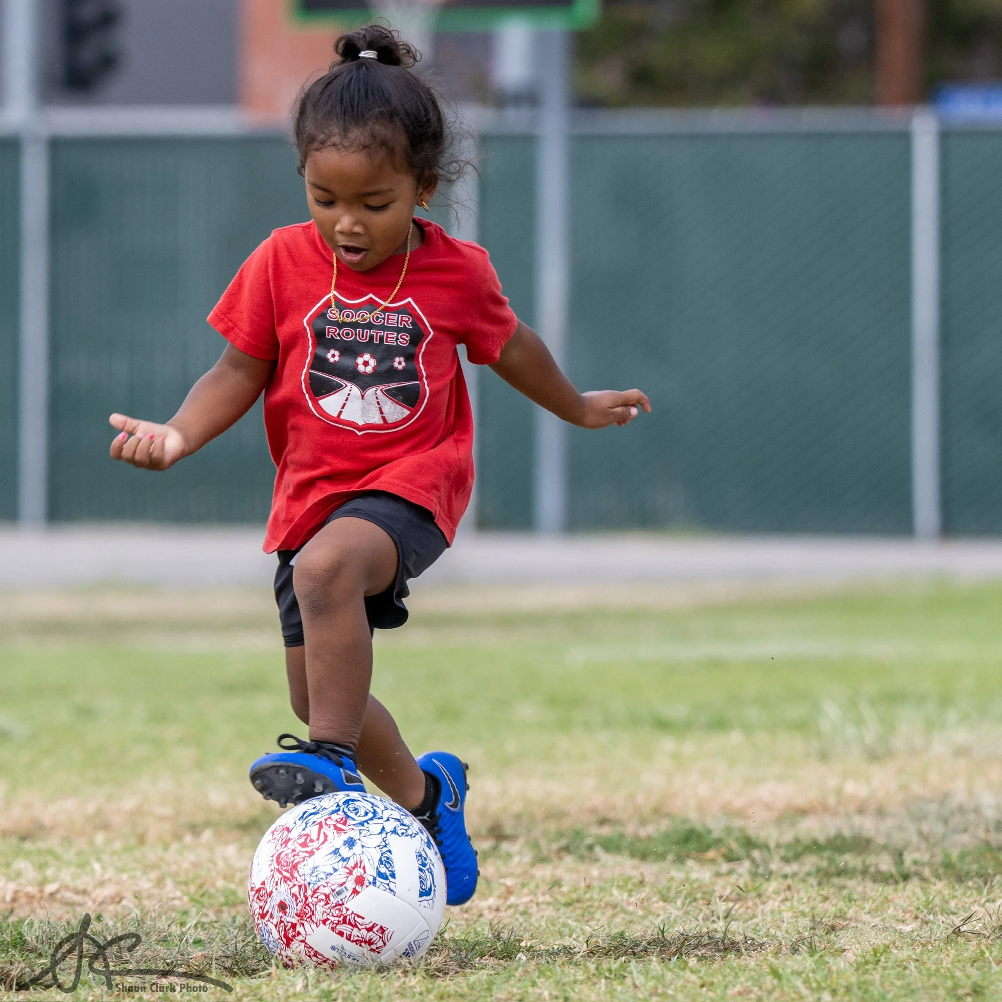 Young girl in a red soccer shirt and blue cleats dribbling a soccer ball on a grassy field