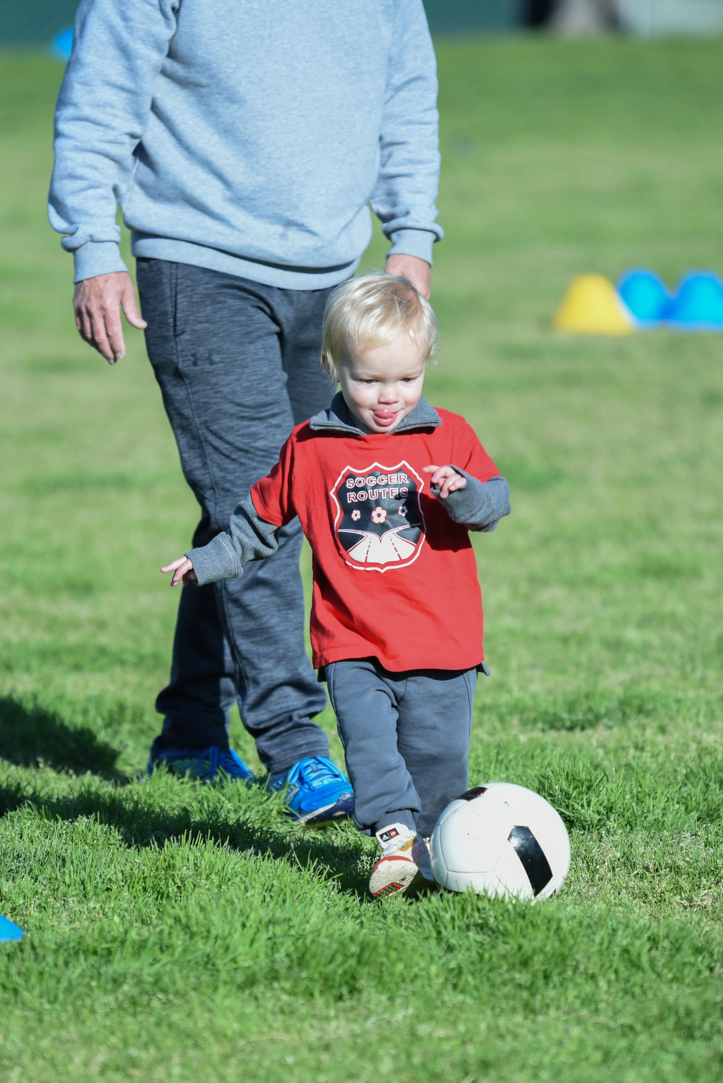 Toddler in a red soccer shirt standing with an adult beside a soccer ball on a grassy field