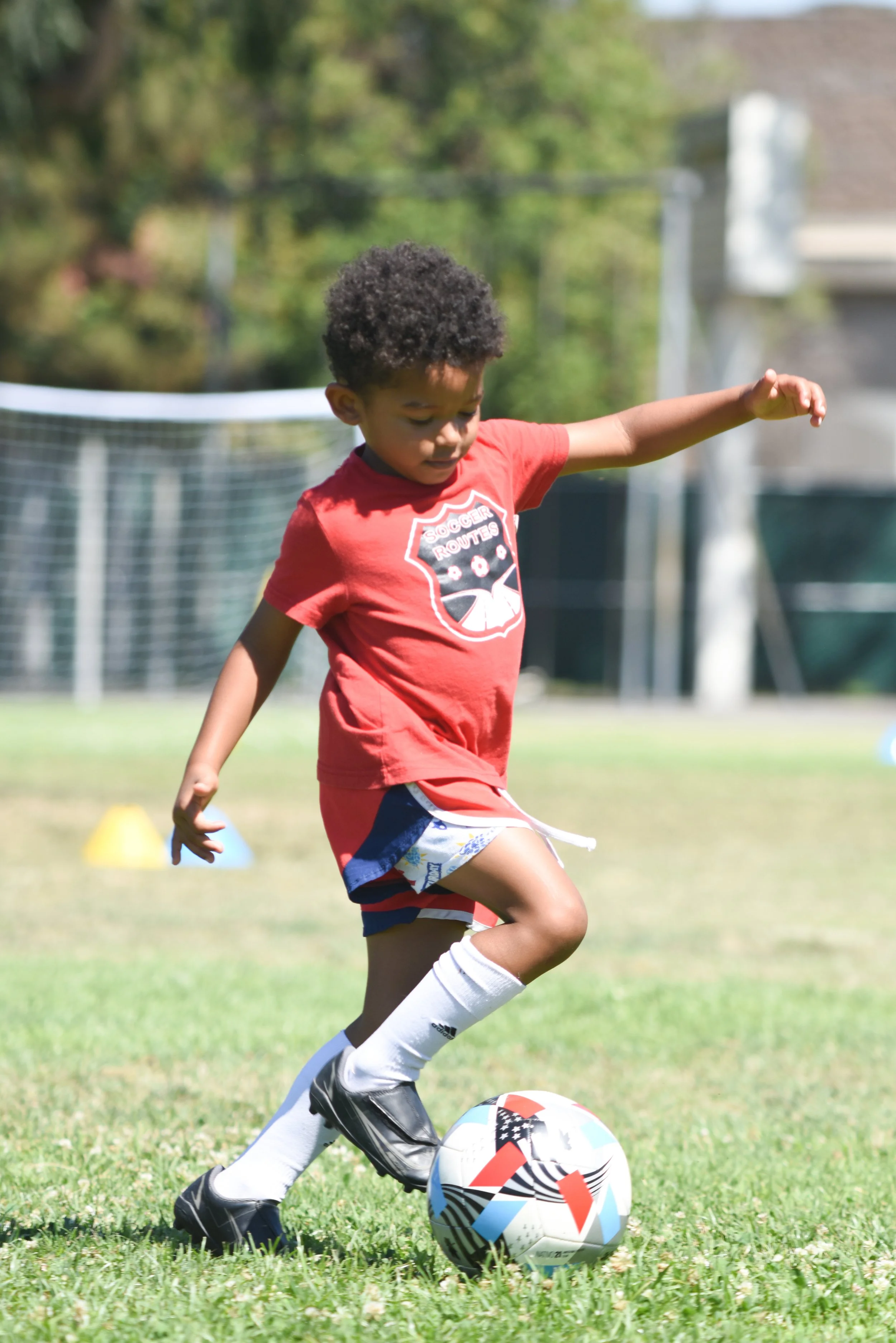 Young child in a red soccer jersey kicking a soccer ball on a grassy field