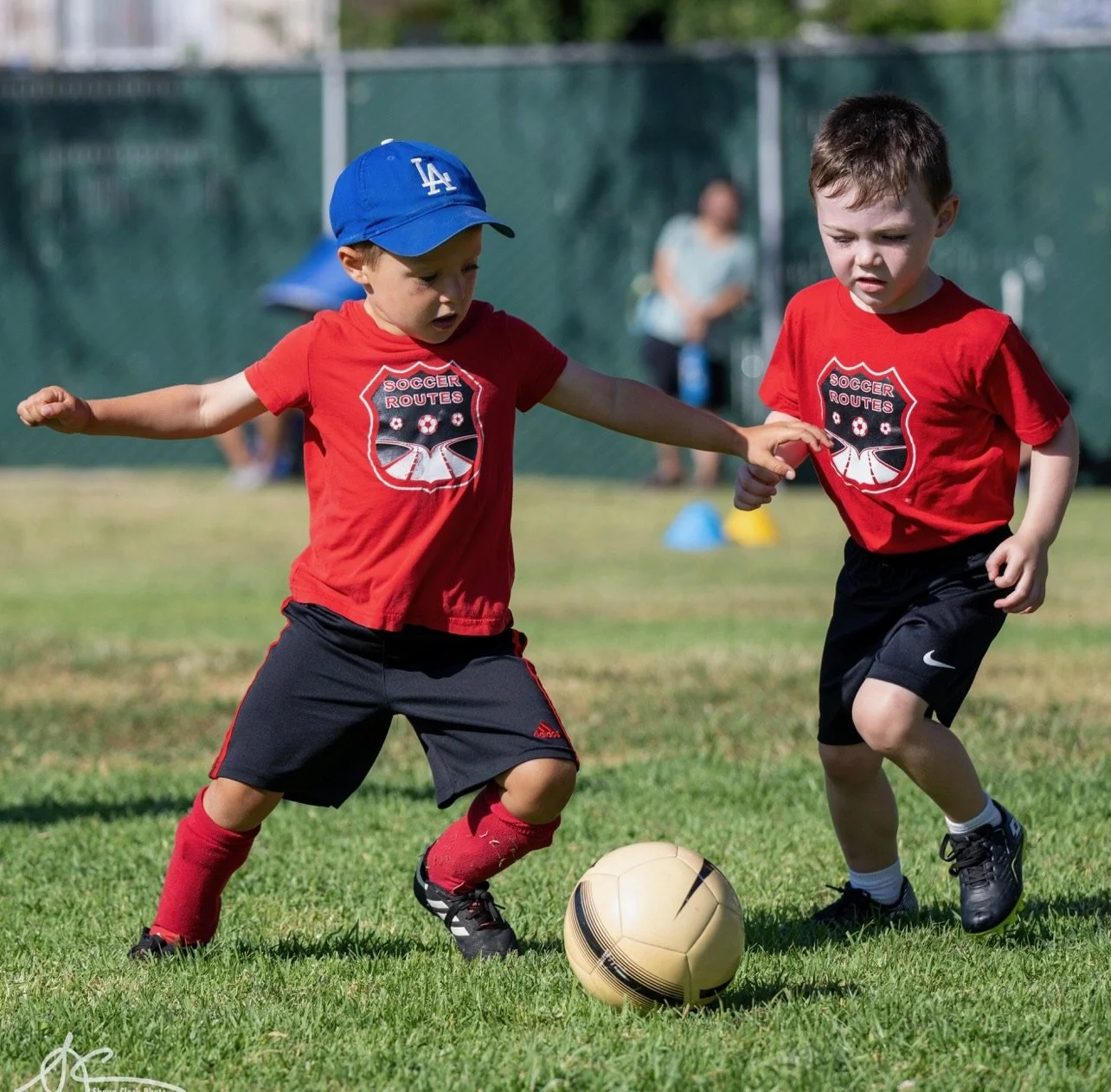 Two young boys in red soccer jerseys running toward a soccer ball on a grassy field.