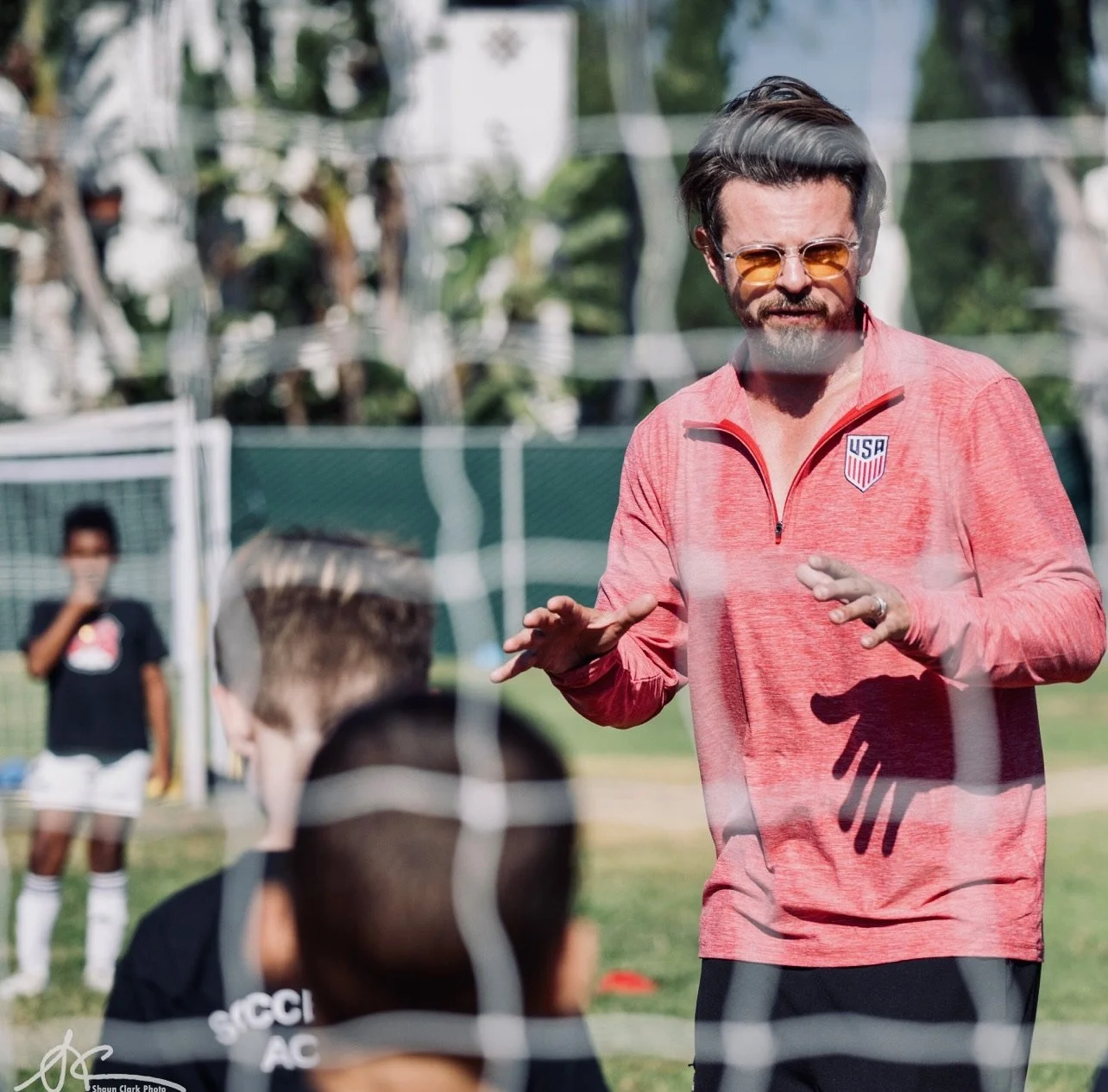 Man in sunglasses and a USA soccer jacket coaching players on an outdoor soccer field