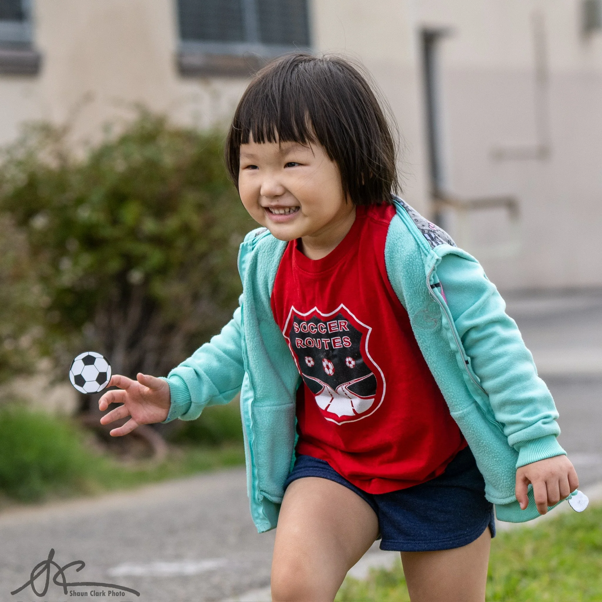 Smiling child in a red soccer shirt and turquoise jacket playing outdoors with a soccer ball