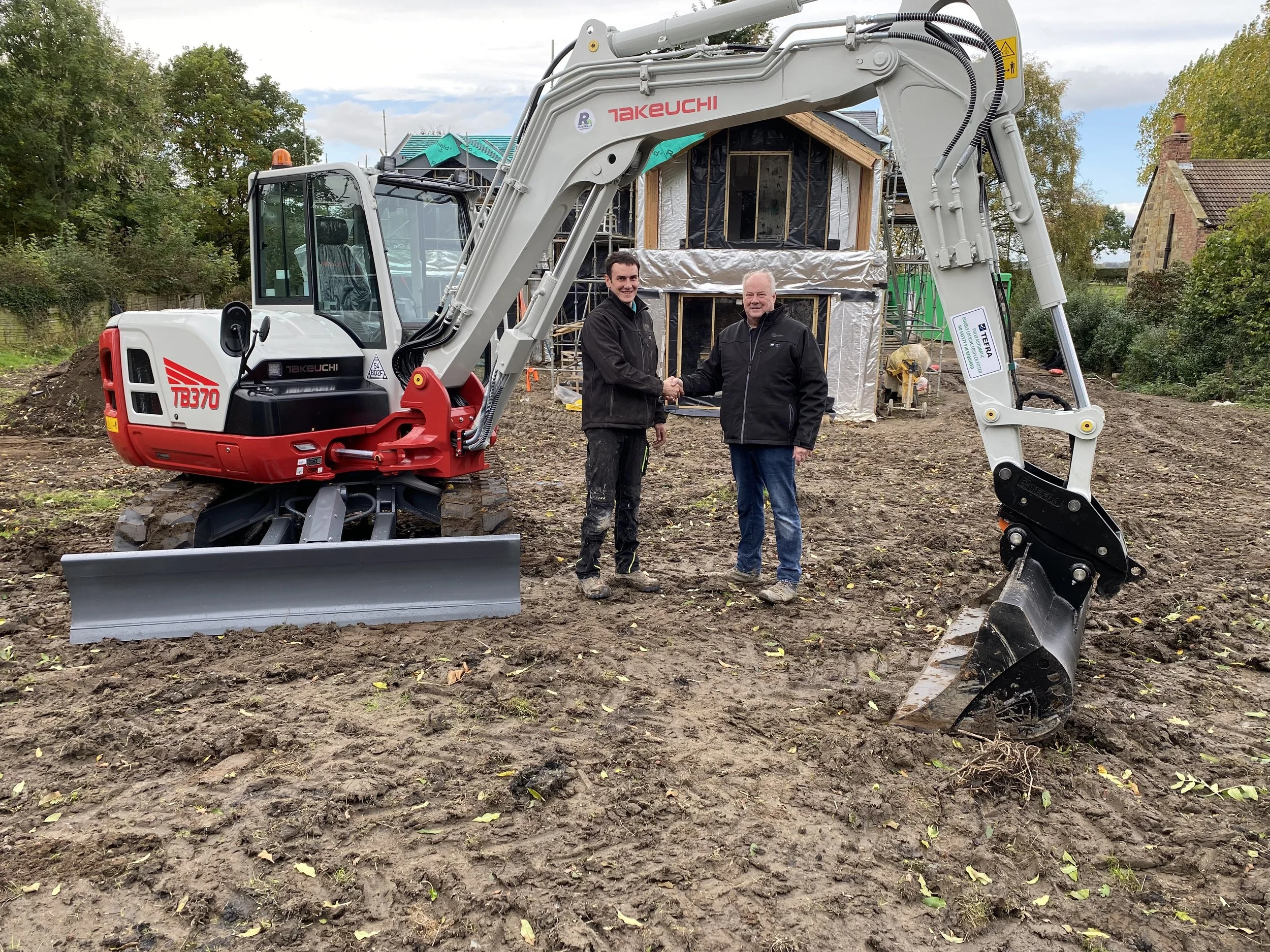 Two men shake hands beside a Takeuchi TB370 excavator at a muddy residential construction site.
