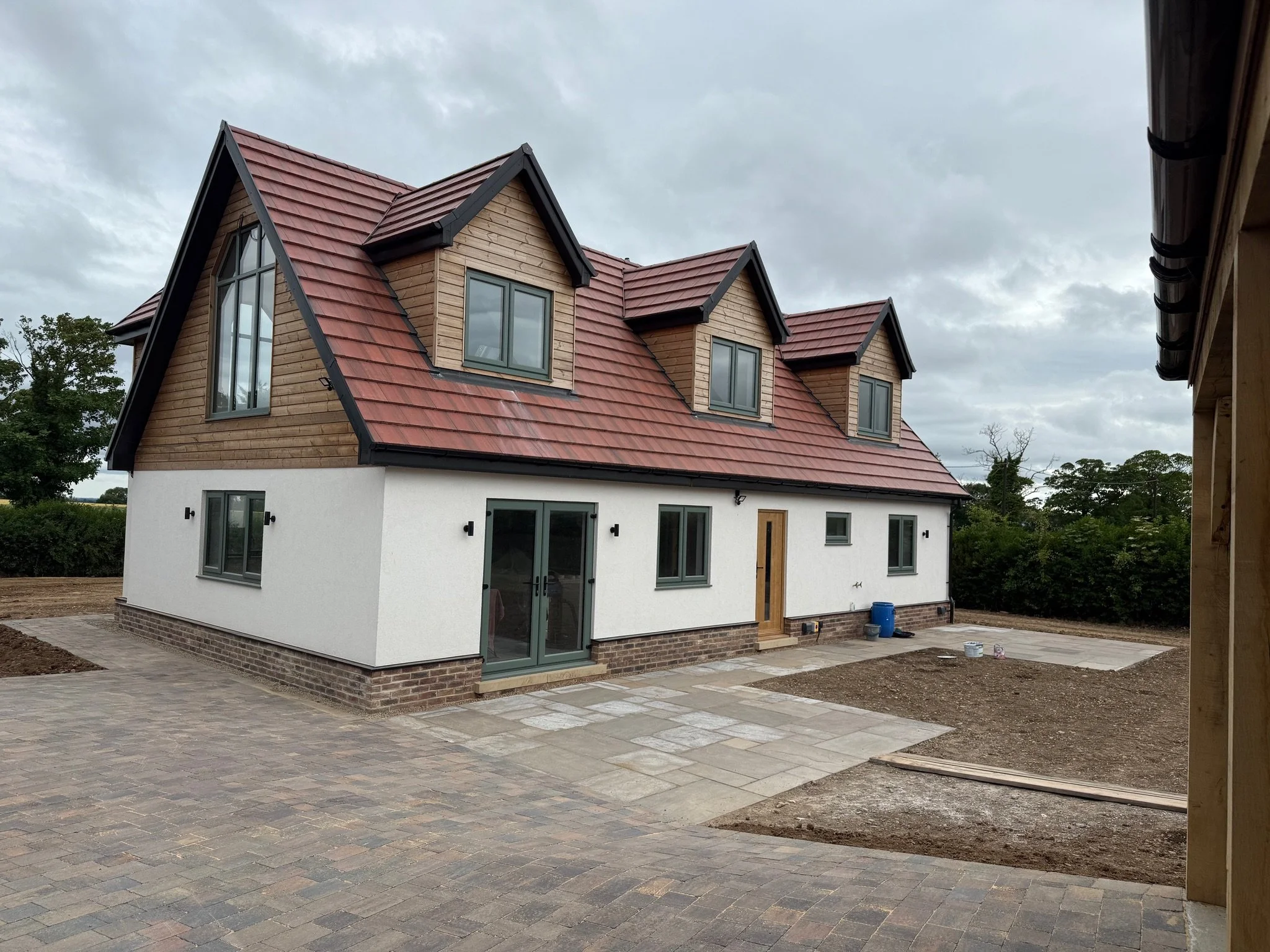 Newly built countryside house with wood siding, white stucco walls, red metal roof, dormer windows, and paved courtyard