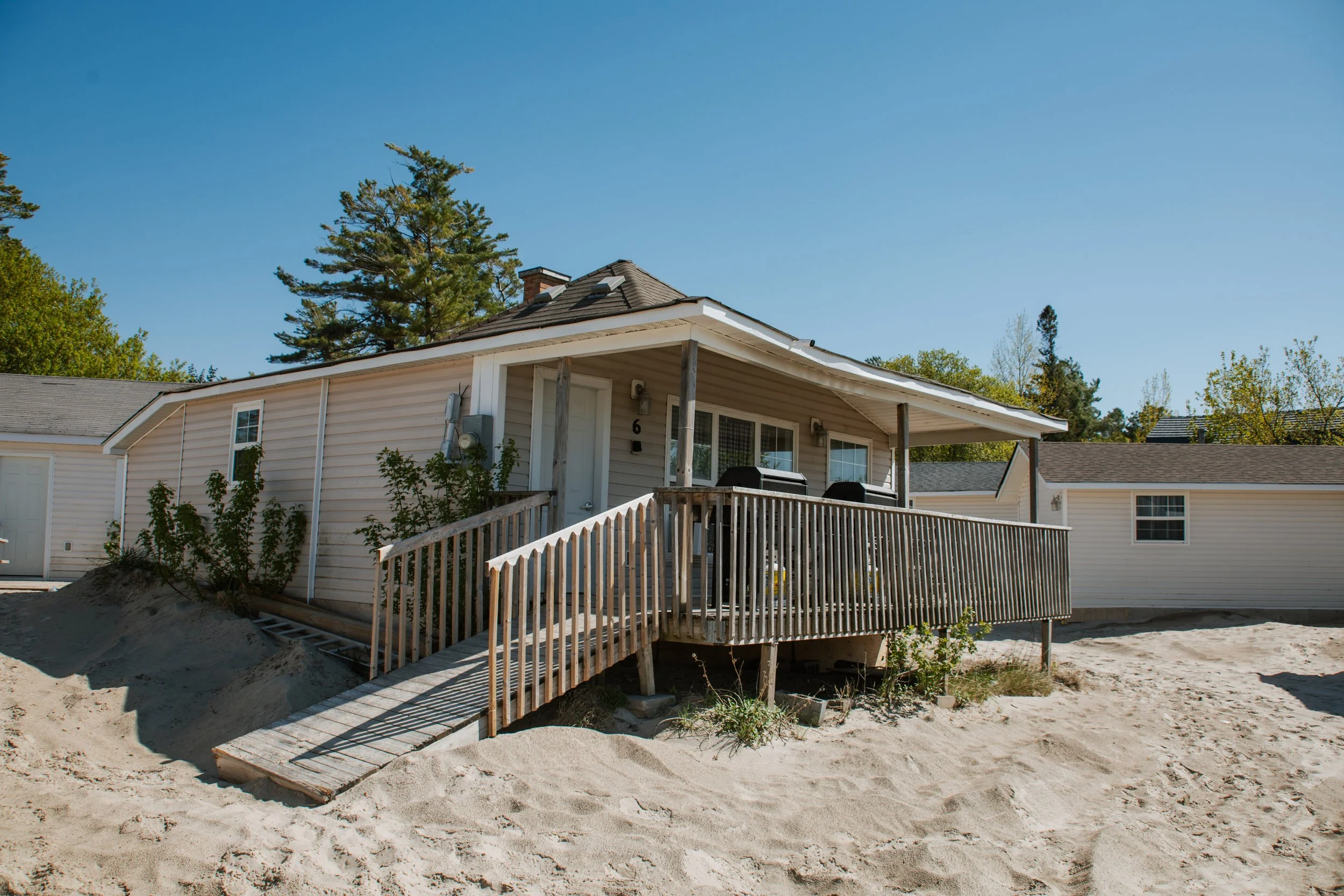 Light-colored beach cottage with a wooden accessibility ramp, porch, grills, trees, and sandy ground under a clear blue sky.