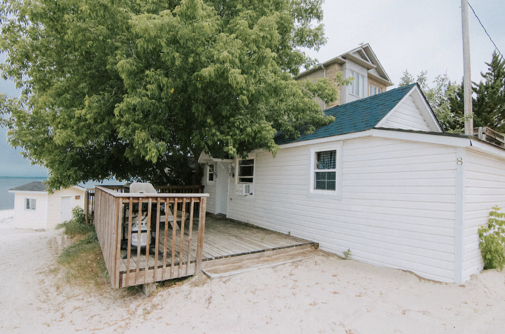 White beach cottage with a teal roof, wooden deck, large shade tree, and lakeshore in the background.
