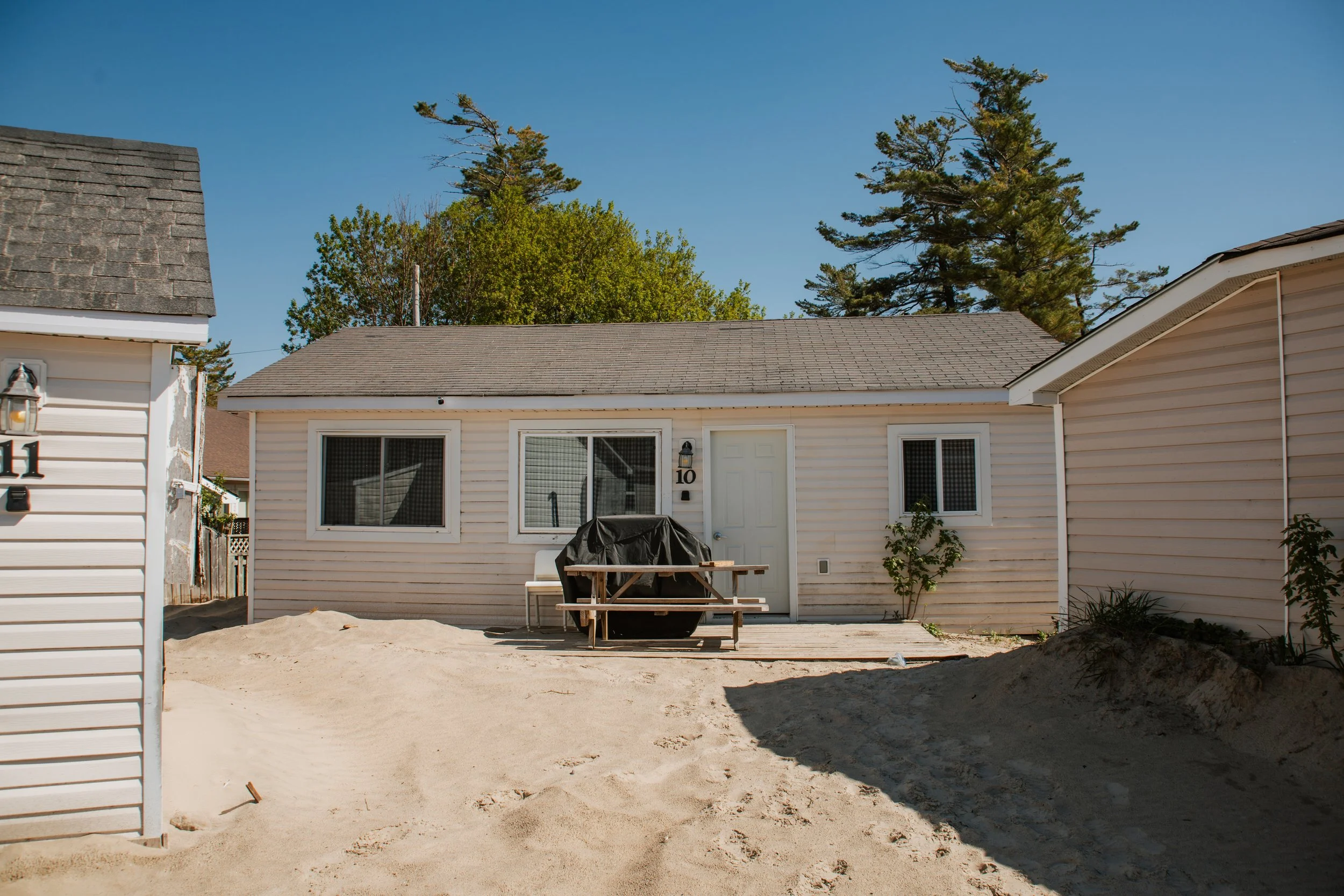 Small beach cottage with a picnic table, sand-covered yard, and pine trees under a clear blue sky