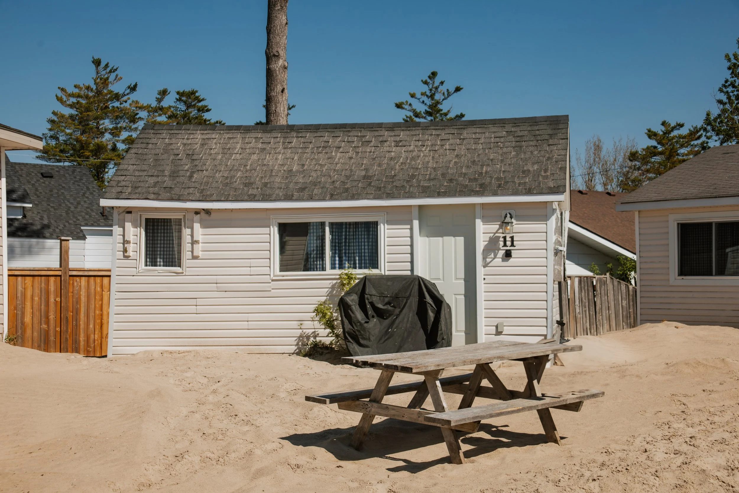 Small beach cottage with a picnic table on sandy ground, surrounded by neighboring cabins and pine trees