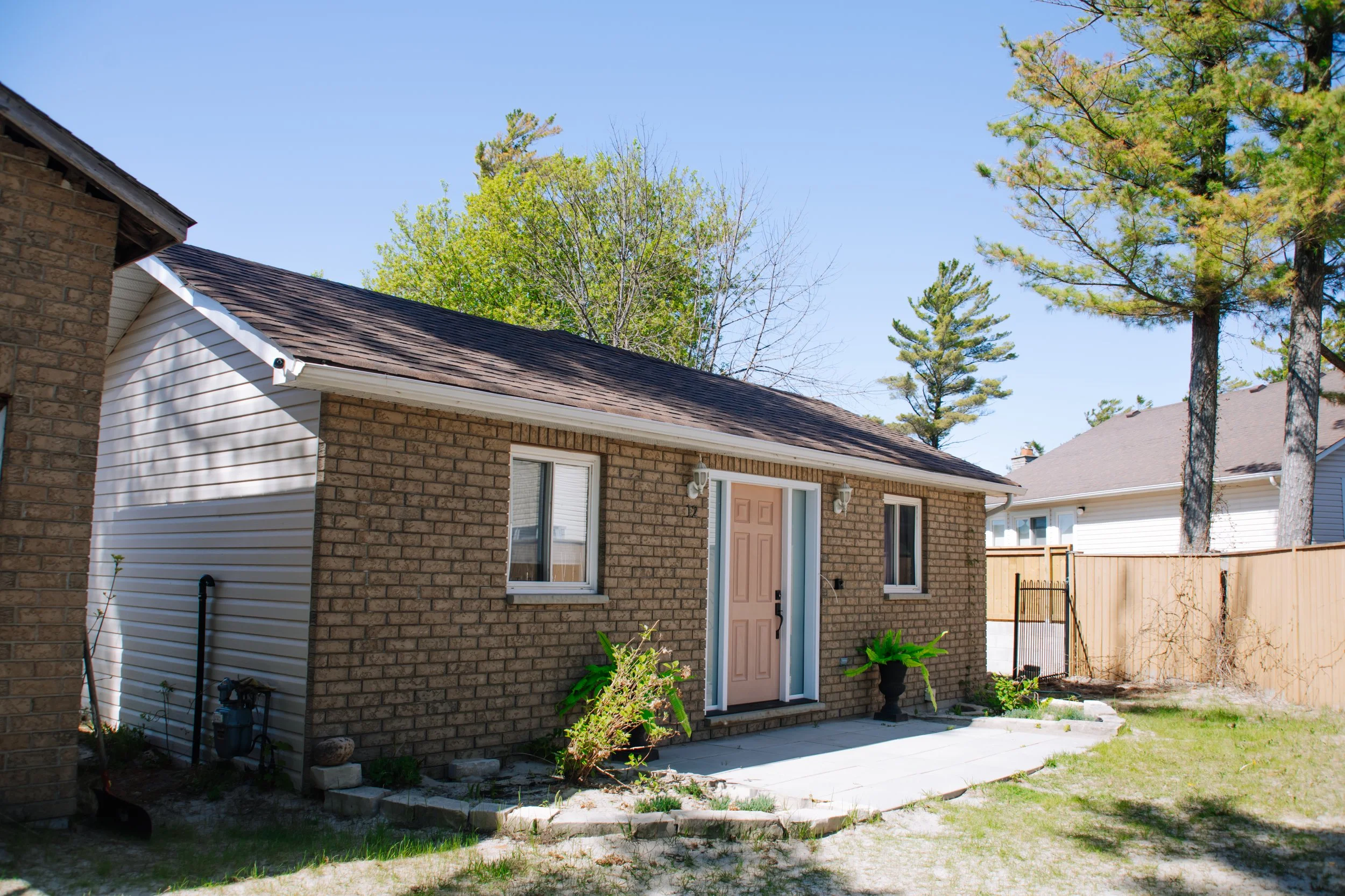 Single-story brick suburban house with a shingled roof, front door, windows, landscaped walkway, and fenced backyard under a clear blue sky