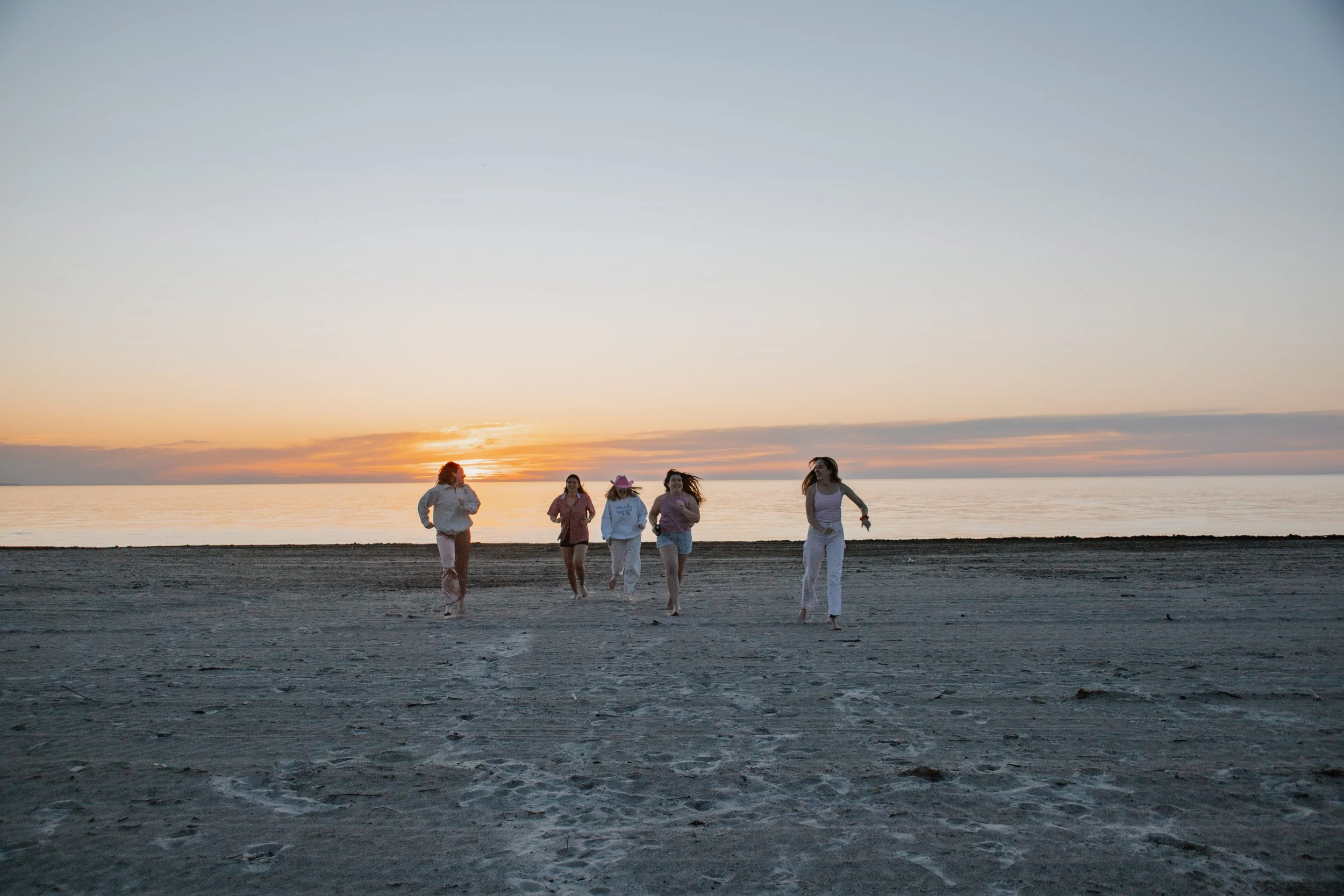Five friends running on a sandy beach at sunset beside calm water