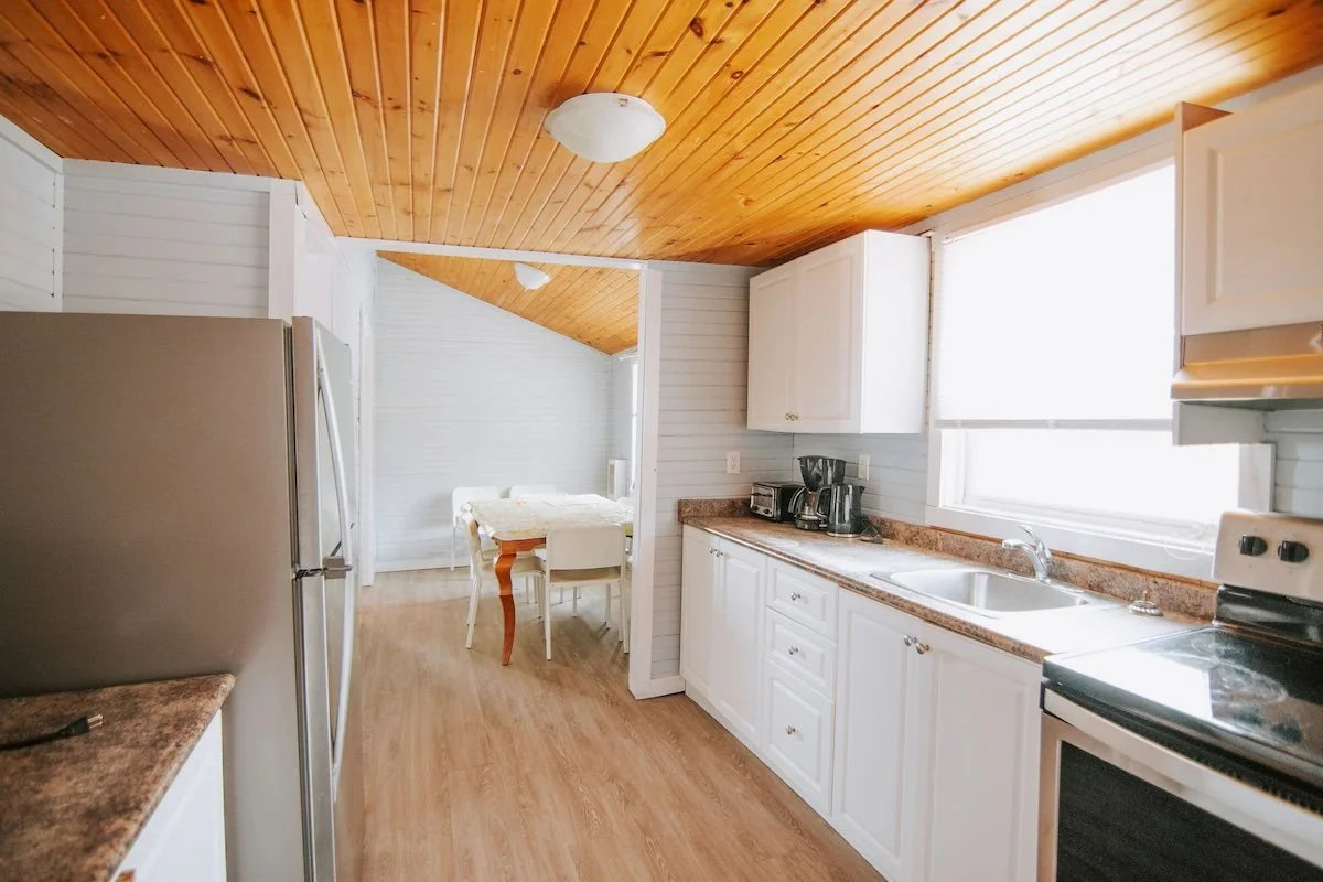 Bright white kitchen with wood-paneled ceiling, stainless steel refrigerator, stove, sink, cabinets, and adjoining dining area